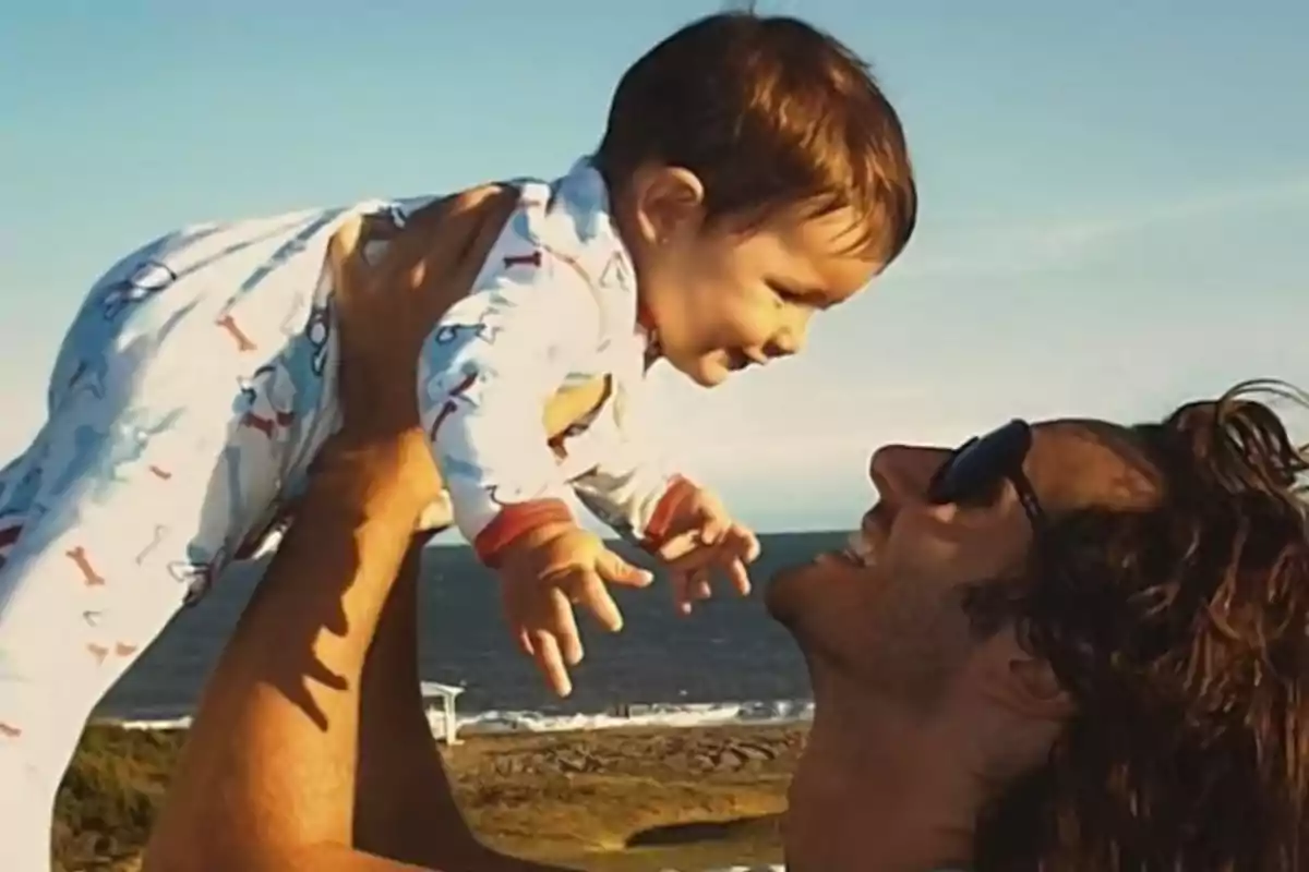 A man holds a baby in the air as they both smile on a sunny day by the sea. A man holds a baby in the air as they both smile on a sunny day by the sea.