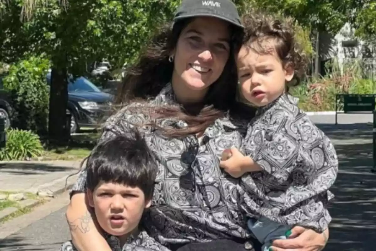Una persona sonriente con dos niños pequeños, todos vistiendo camisas con un patrón similar, posan al aire libre en un día soleado. Una persona sonriente con dos niños pequeños, todos vistiendo camisas con un patrón similar, posan al aire libre en un día soleado.