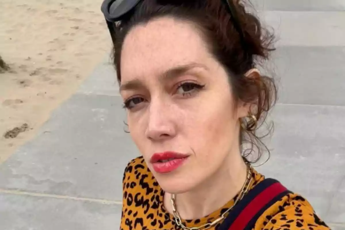 A person with dark hair and sunglasses on their head, wearing an animal print blouse, is on a beach with sand and pavement.