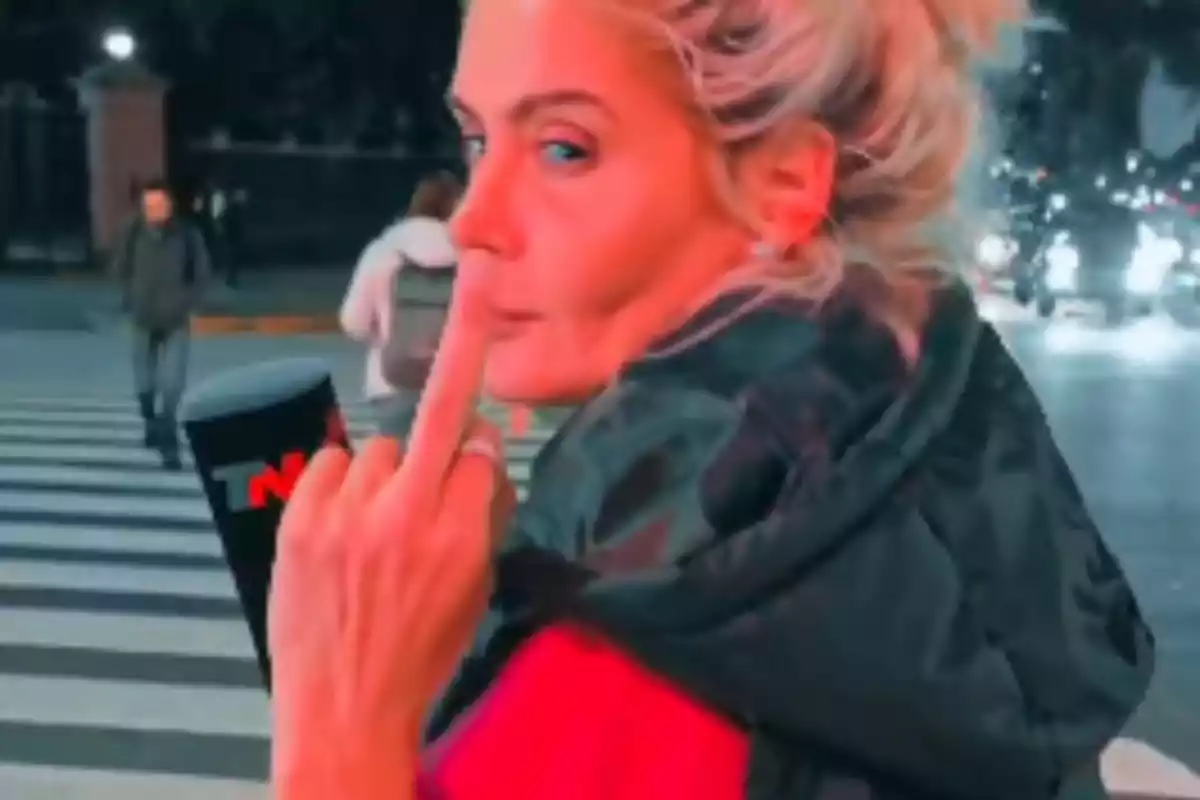 A person with blonde hair tied up is at a crosswalk at night, looking back while raising a finger. They are wearing a dark jacket and holding a microphone with the "TN" logo. In the background, there are car lights and people walking.