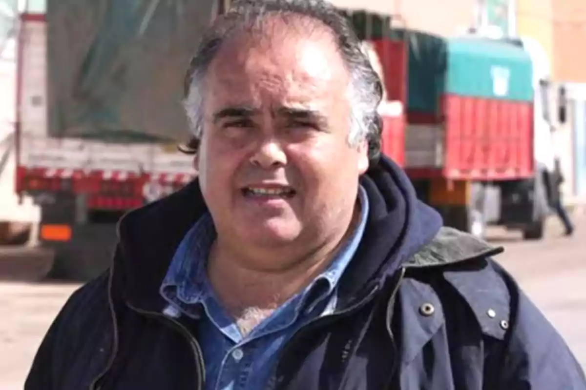 A dark-haired man in a blue jacket is standing outdoors with trucks in the background.