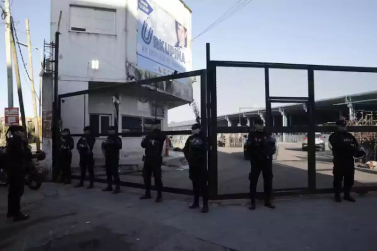 A group of police officers is lined up in front of an entrance with a metal gate, next to a white building with a billboard. A group of police officers is lined up in front of an entrance with a metal gate, next to a white building with a billboard.