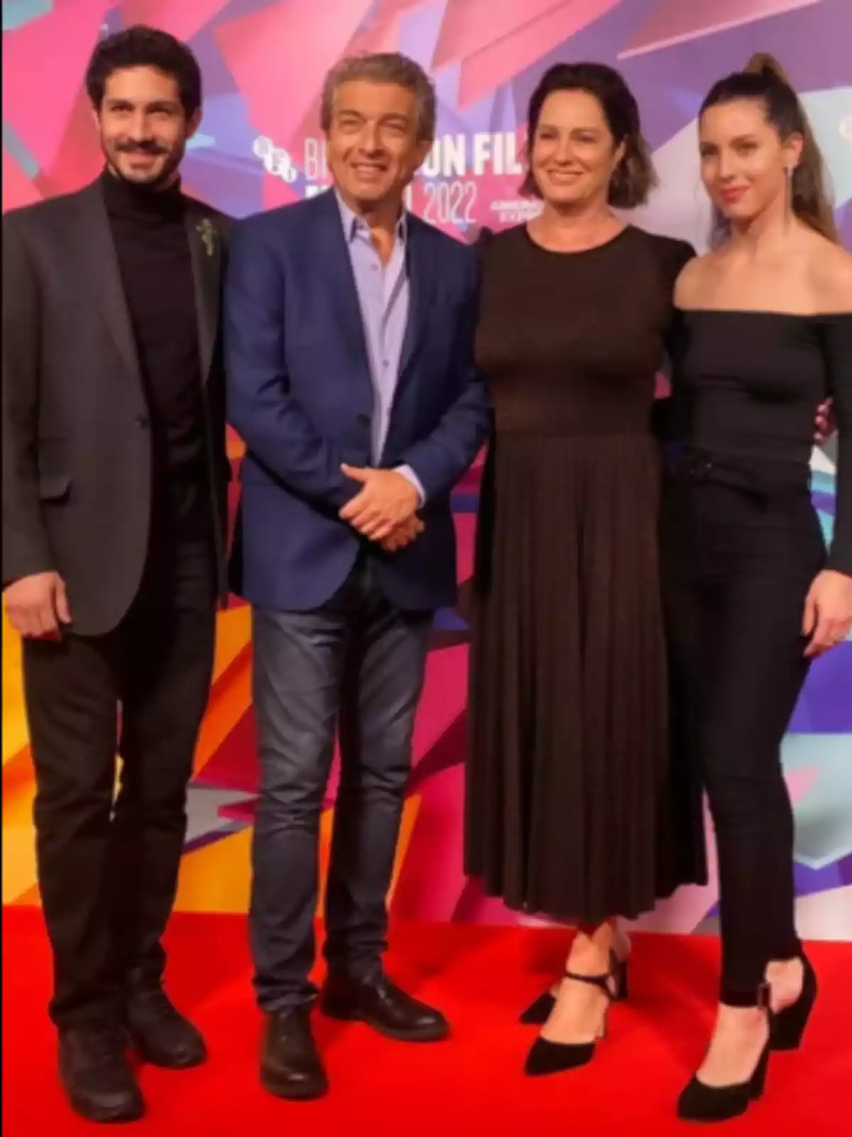 Four people pose together on a red carpet with a colorful background. Four people pose together on a red carpet with a colorful background.
