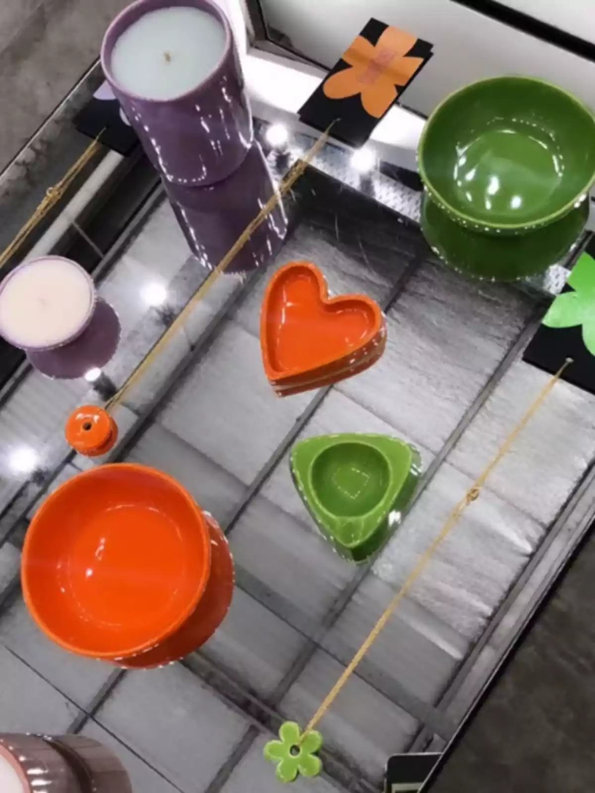 A table with a mirrored surface reflects various colorful objects, including candles in purple holders, ceramic plates in heart and triangle shapes in orange and green, and necklaces with flower pendants. A table with a mirrored surface reflects various colorful objects, including candles in purple holders, ceramic plates in heart and triangle shapes in orange and green, and necklaces with flower pendants.