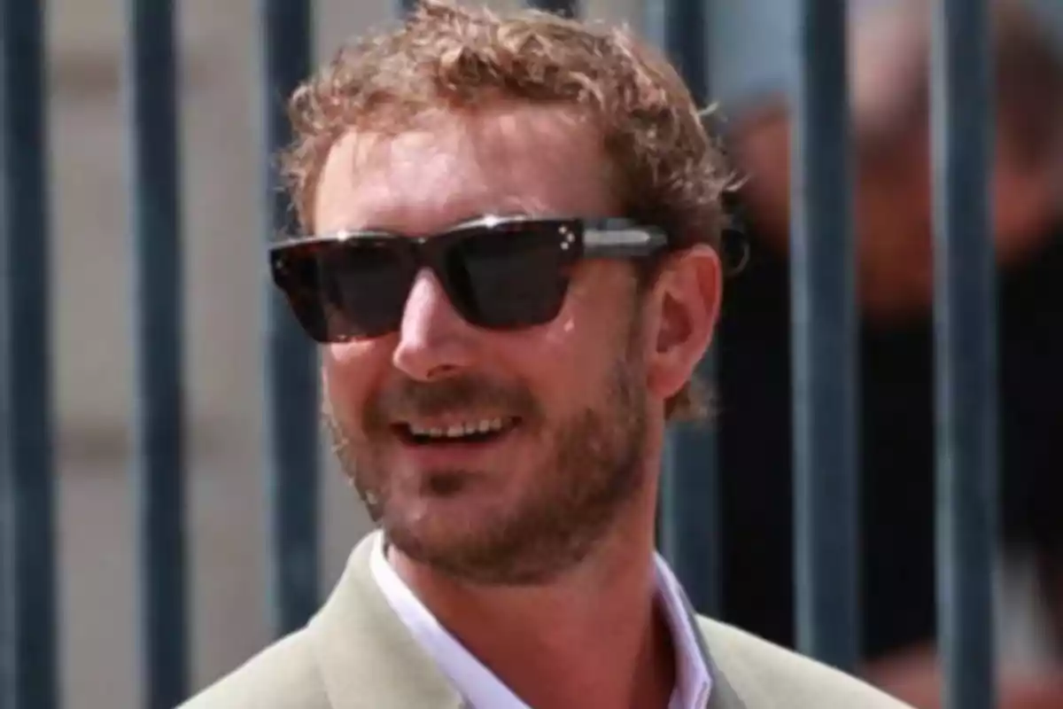 A man with a beard and sunglasses smiles while wearing a light suit.