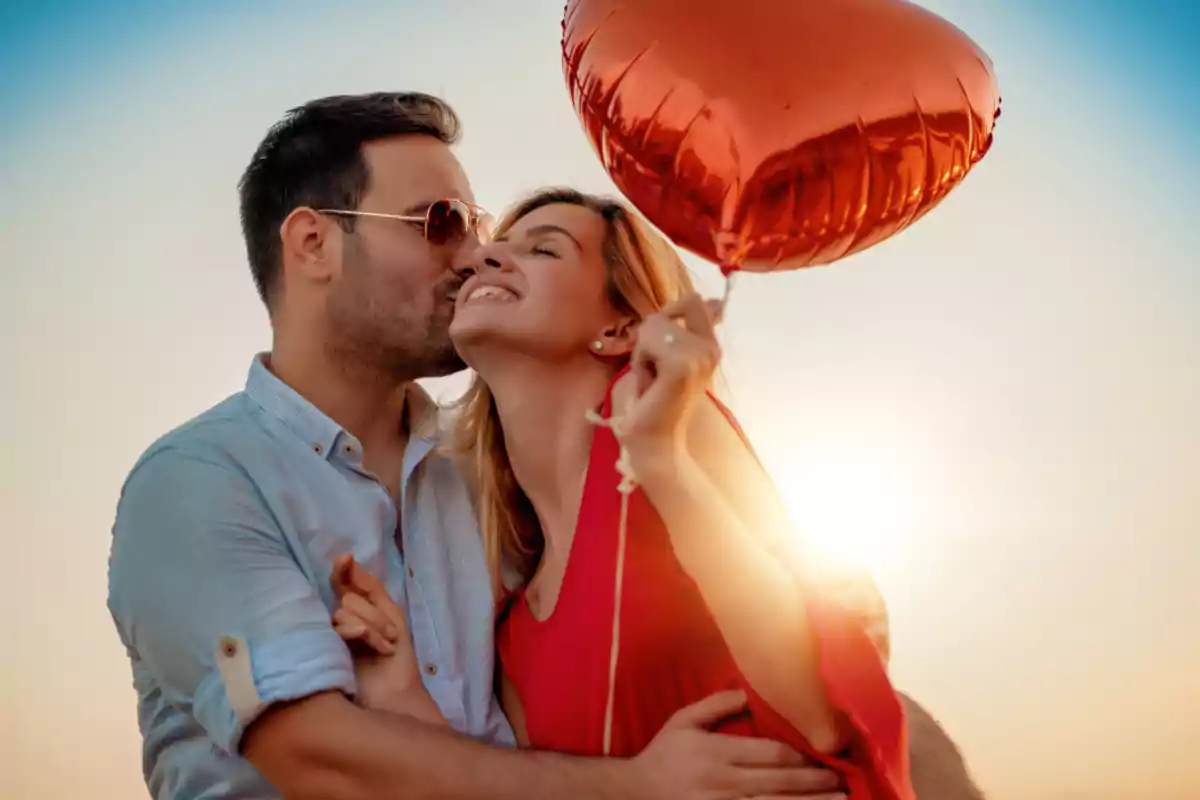 Una pareja sonriente se abraza mientras el hombre besa a la mujer en la mejilla, ella sostiene un globo rojo en forma de corazón. Una pareja sonriente se abraza mientras el hombre besa a la mujer en la mejilla, ella sostiene un globo rojo en forma de corazón.