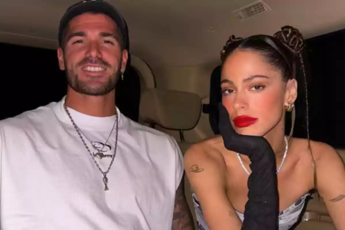 Two people pose sitting in the back seat of a car, one of them smiling and the other resting their face on a gloved hand; both look well-dressed and are looking at the camera.