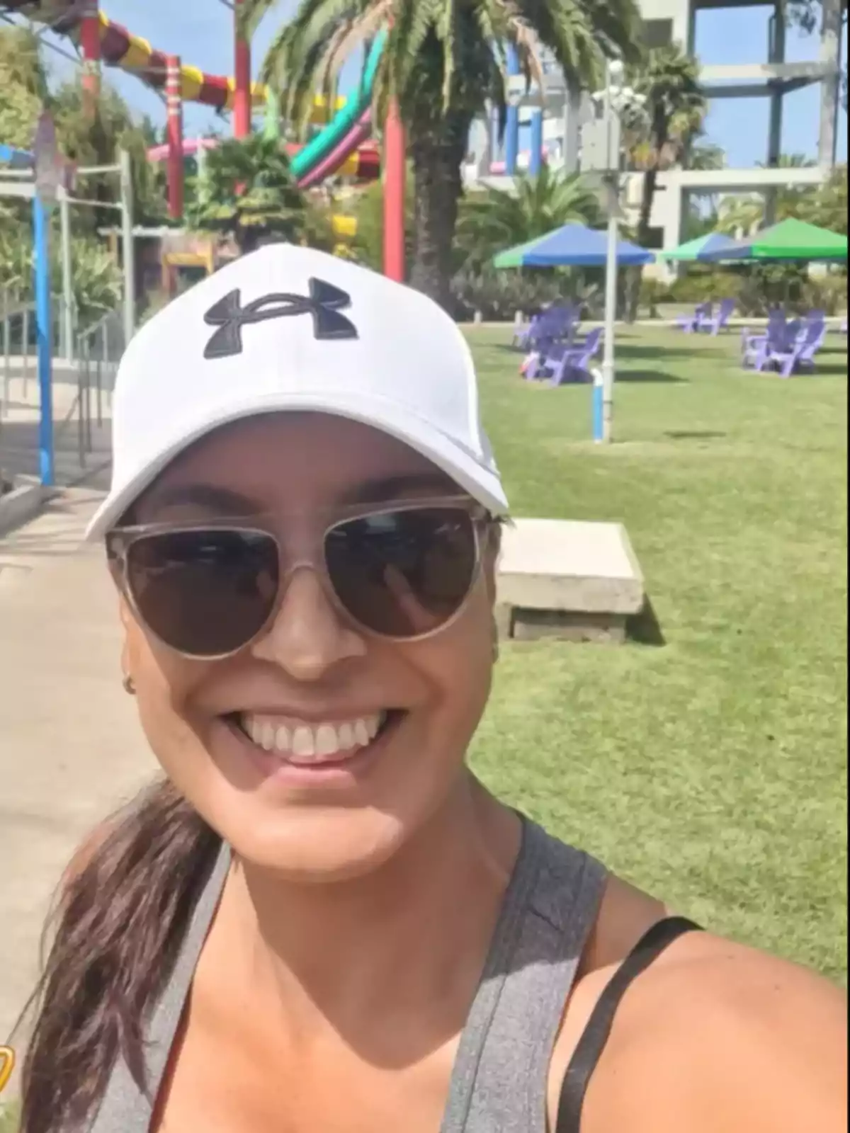 Mujer sonriente con gorra blanca y gafas de sol tomándose una selfie en un parque acuático con toboganes de colores y palmeras al fondo Mujer sonriente con gorra blanca y gafas de sol tomándose una selfie en un parque acuático con toboganes de colores y palmeras al fondo