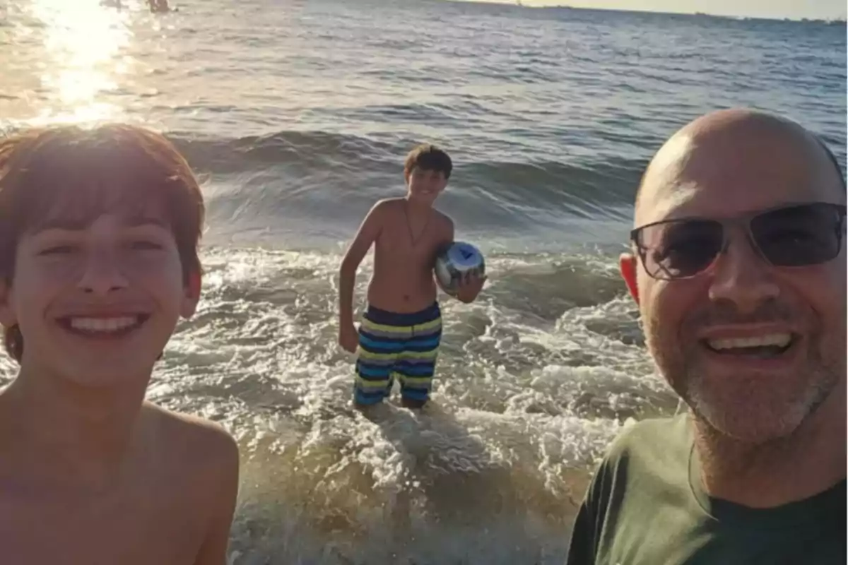 Tres personas sonríen y disfrutan en la orilla del mar al atardecer, una de ellas sostiene un balón y el sol brilla sobre el agua. Tres personas sonríen y disfrutan en la orilla del mar al atardecer, una de ellas sostiene un balón y el sol brilla sobre el agua.