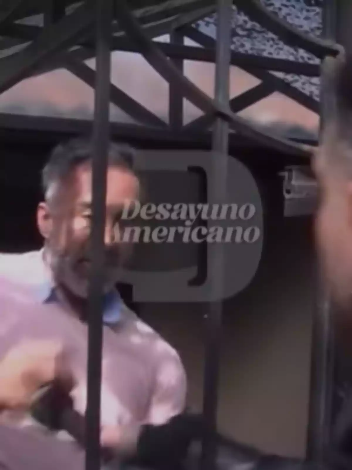 A gray-haired, bearded man is behind a gate while interacting with another person; the Desayuno Americano logo appears in the center of the image. A gray-haired, bearded man is behind a gate while interacting with another person; the Desayuno Americano logo appears in the center of the image.