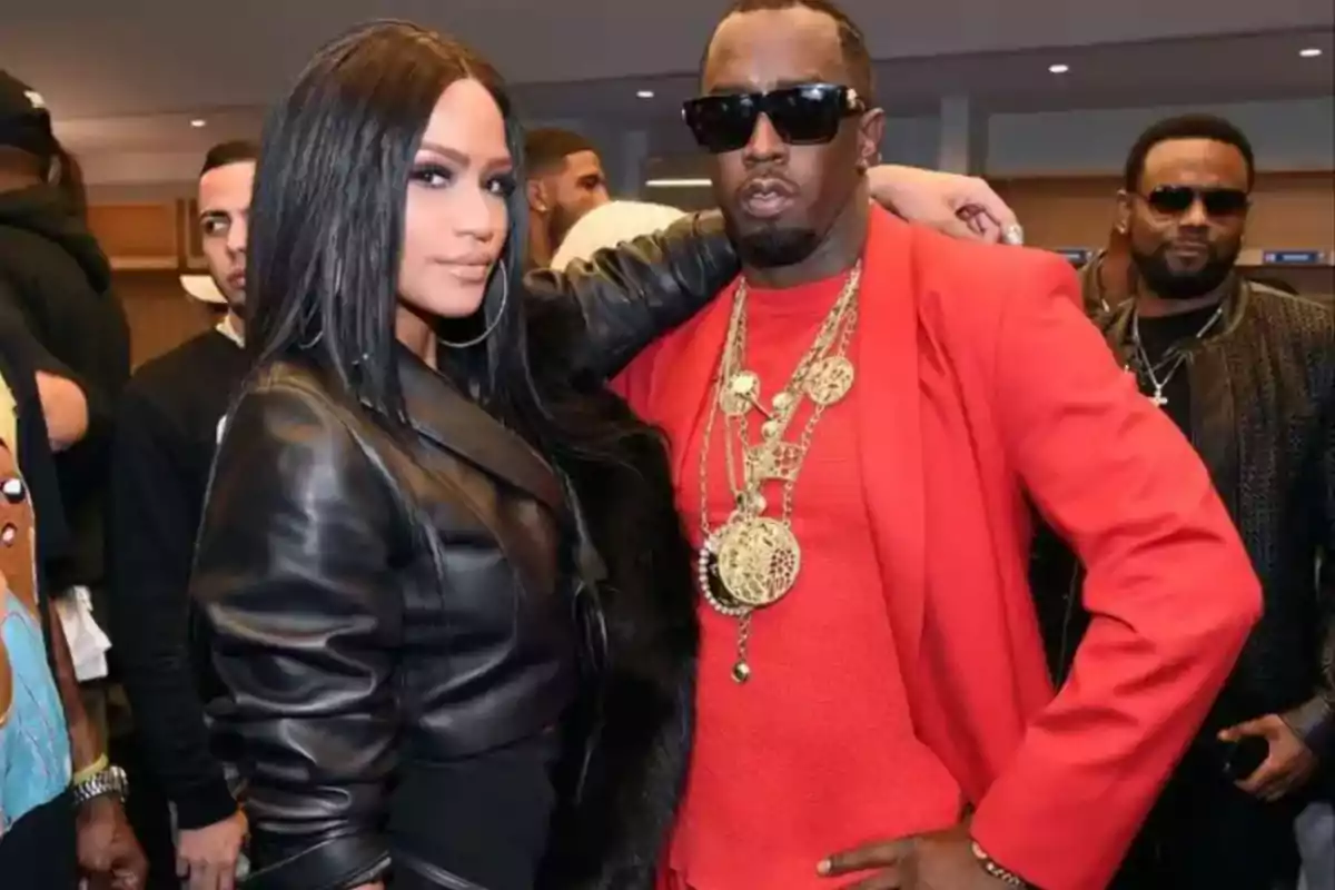 A woman in a black leather jacket and a man wearing dark glasses and a red jacket pose together at an event surrounded by other people. A woman in a black leather jacket and a man wearing dark glasses and a red jacket pose together at an event surrounded by other people.