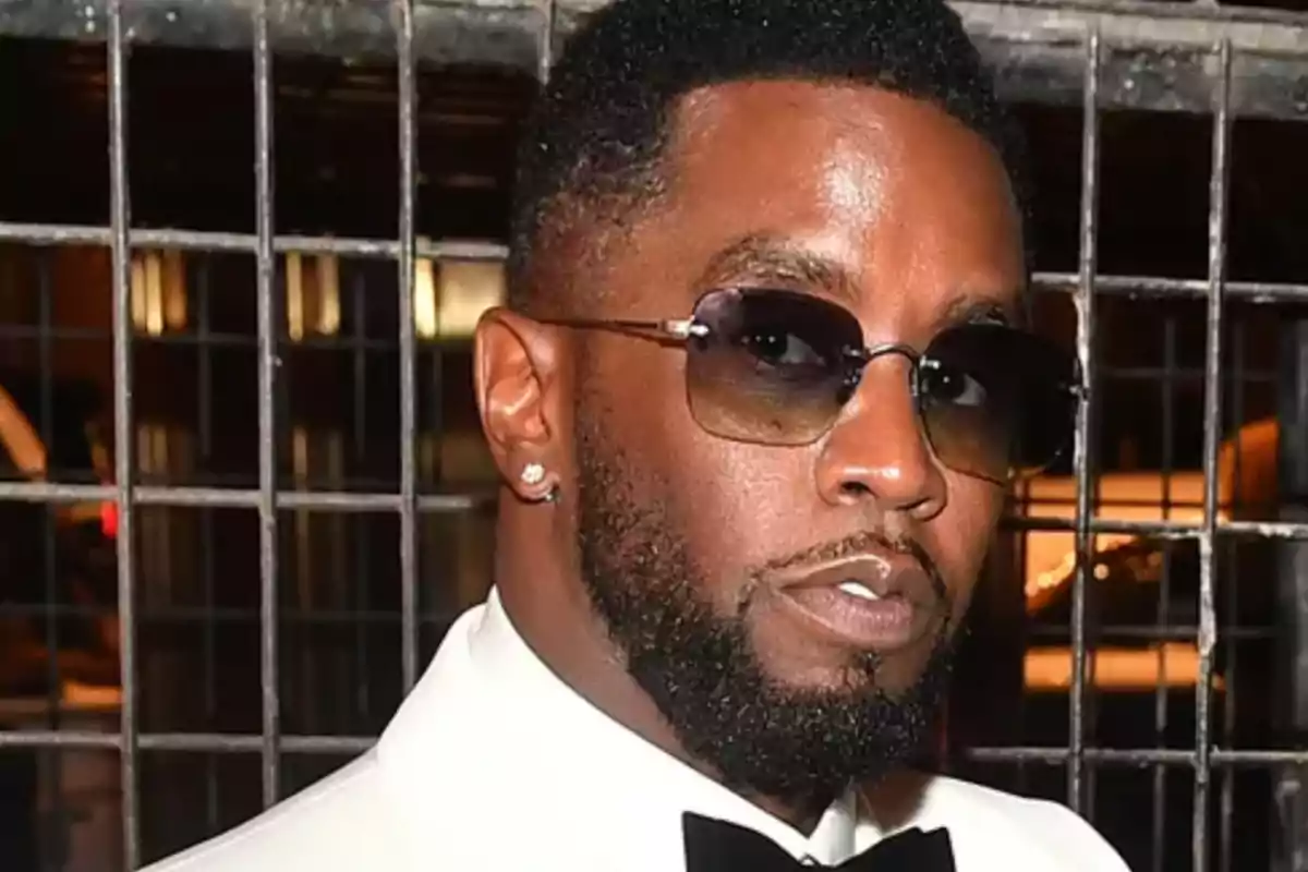 Man with sunglasses, beard, and mustache, dressed in a white tuxedo and black bow tie, posing in front of a metal fence.