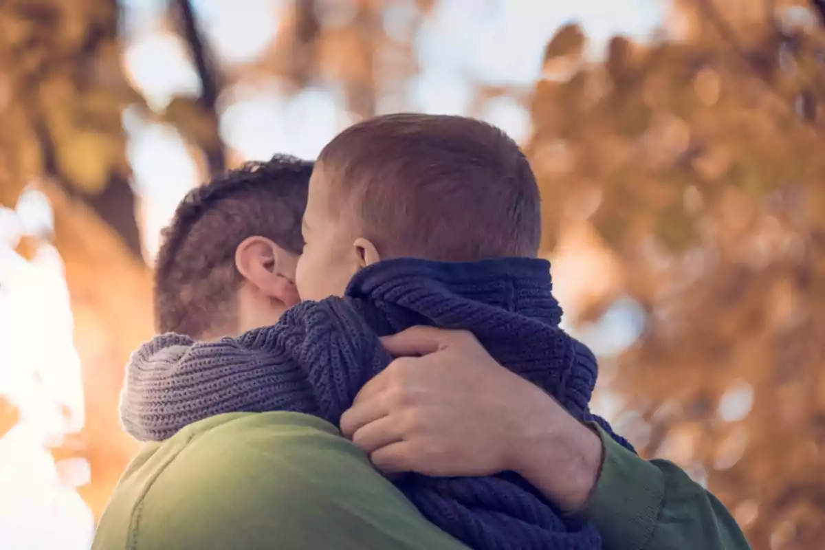 Un hombre abraza a un niño pequeño envuelto en una bufanda azul con un fondo de árboles otoñales desenfocados.