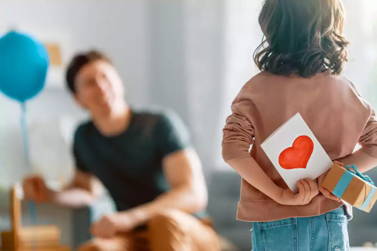 Niña sosteniendo una tarjeta con un corazón rojo y un regalo detrás de la espalda mientras un hombre la observa sonriente en el fondo Niña sosteniendo una tarjeta con un corazón rojo y un regalo detrás de la espalda mientras un hombre la observa sonriente en el fondo