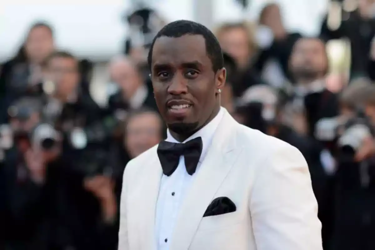 Man in a white tuxedo and black bow tie posing at a formal event with people and cameras in the background Man in a white tuxedo and black bow tie posing at a formal event with people and cameras in the background