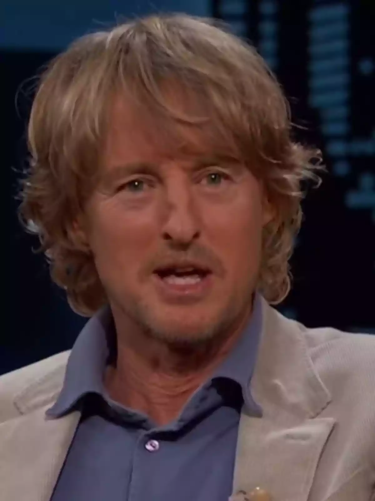Blond, wavy-haired man wearing a beige jacket and blue jersey in a television studio setting Blond, wavy-haired man wearing a beige jacket and blue jersey in a television studio setting