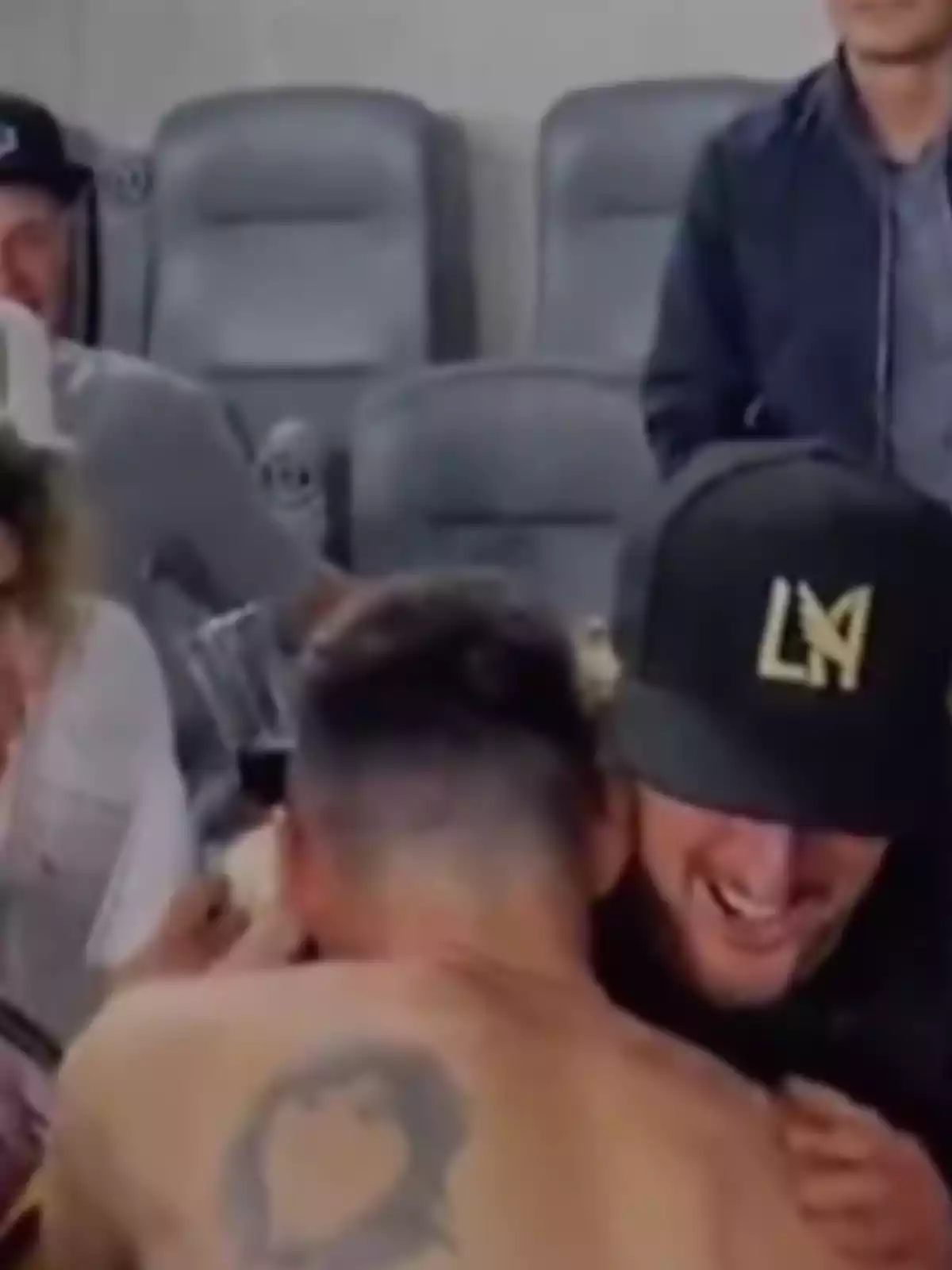Two people hug joyfully in a stadium; one of them is wearing a black cap with the letters LA, and the other is jerseyless, showing a tattoo on their back. Two people hug joyfully in a stadium; one of them is wearing a black cap with the letters LA, and the other is jerseyless, showing a tattoo on their back.