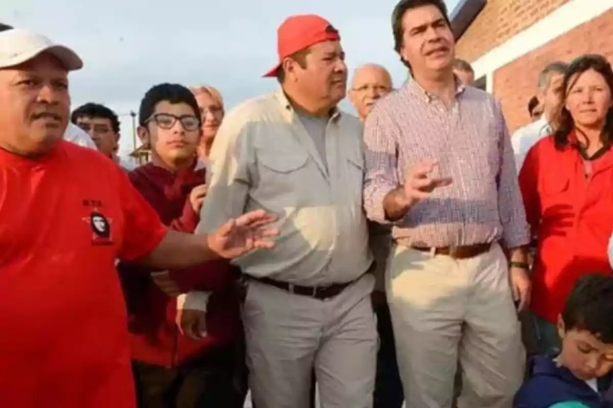 A group of people of different ages is gathered outdoors; some are wearing casual clothes and others are wearing red t-jerseys. Everyone seems to be paying attention to a conversation or activity. A group of people of different ages is gathered outdoors; some are wearing casual clothes and others are wearing red t-jerseys. Everyone seems to be paying attention to a conversation or activity.