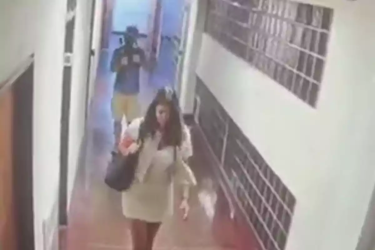 A woman walks down a hallway while a man follows her at a distance.