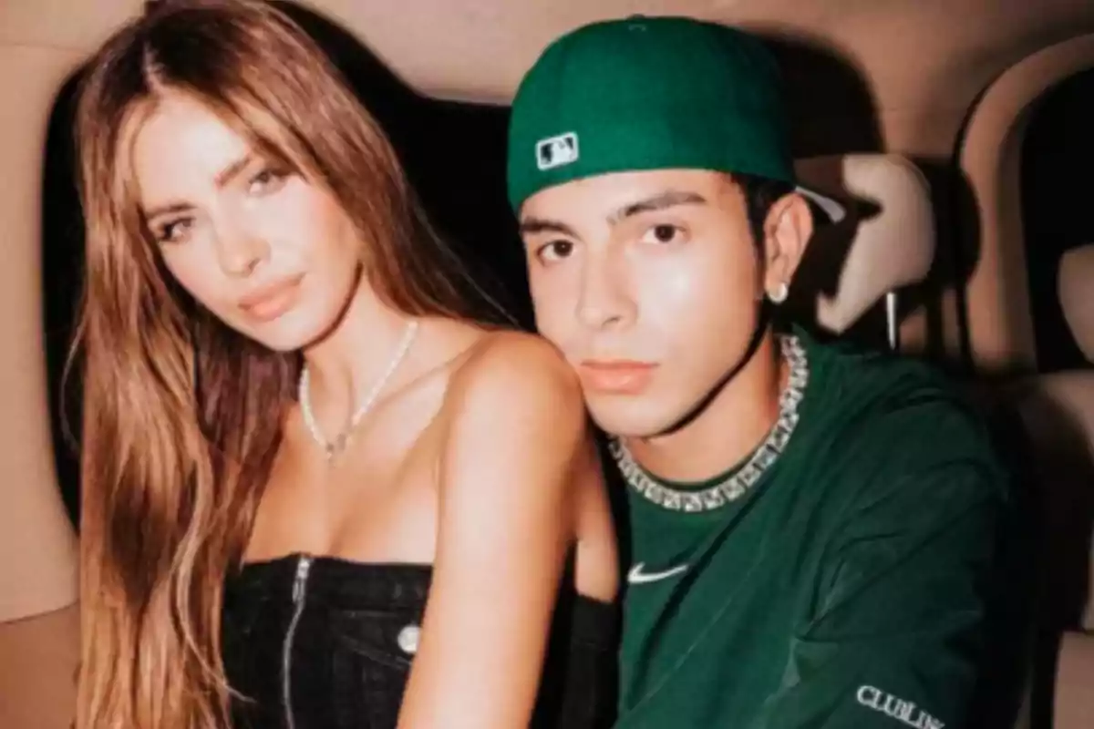 A woman with long hair and a man wearing a green cap pose together sitting inside a car.