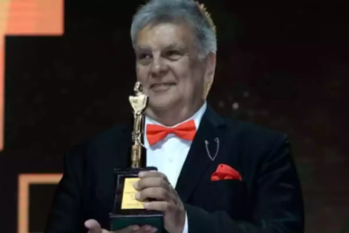 Gray-haired man in a tuxedo and red bow tie holding a golden trophy while smiling on a stage