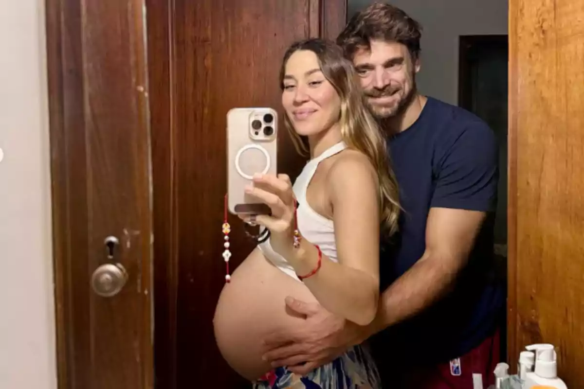 Smiling couple taking a selfie in front of the mirror while the pregnant woman shows her belly and the man hugs her from behind Smiling couple taking a selfie in front of the mirror while the pregnant woman shows her belly and the man hugs her from behind