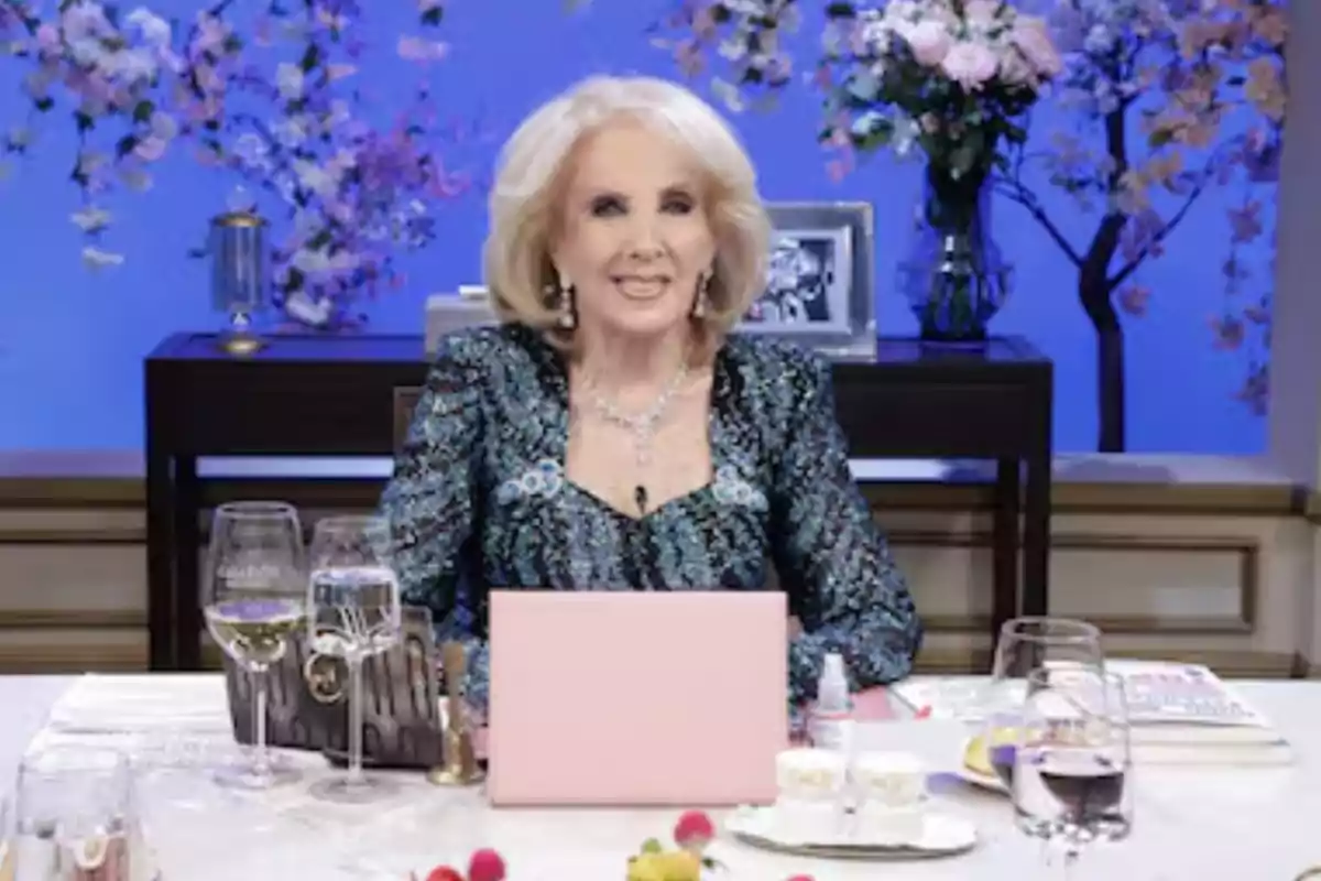 Mujer mayor elegantemente vestida sentada en una mesa decorada con copas, flores y dulces, con un fondo azul y arreglos florales