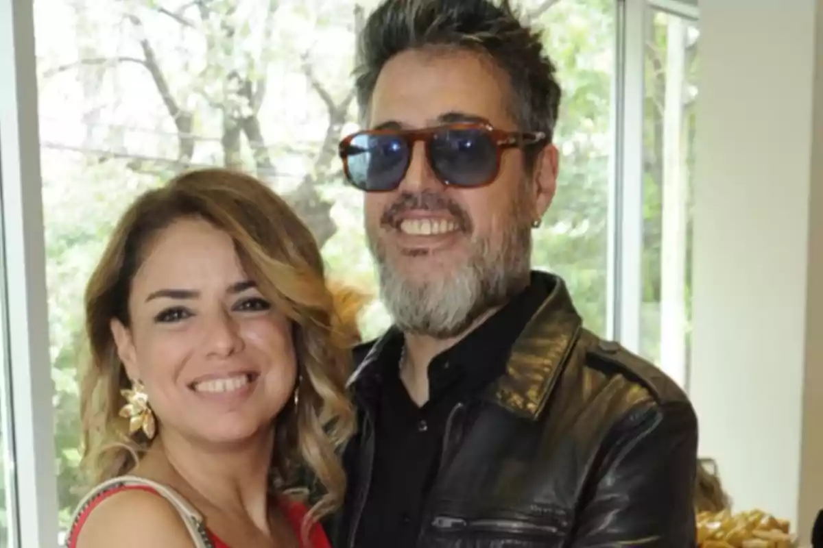 Pareja sonriendo frente a una ventana con fondo de árboles, el hombre lleva gafas oscuras y chaqueta de cuero negra, la mujer usa un vestido rojo y aretes dorados. Pareja sonriendo frente a una ventana con fondo de árboles, el hombre lleva gafas oscuras y chaqueta de cuero negra, la mujer usa un vestido rojo y aretes dorados.