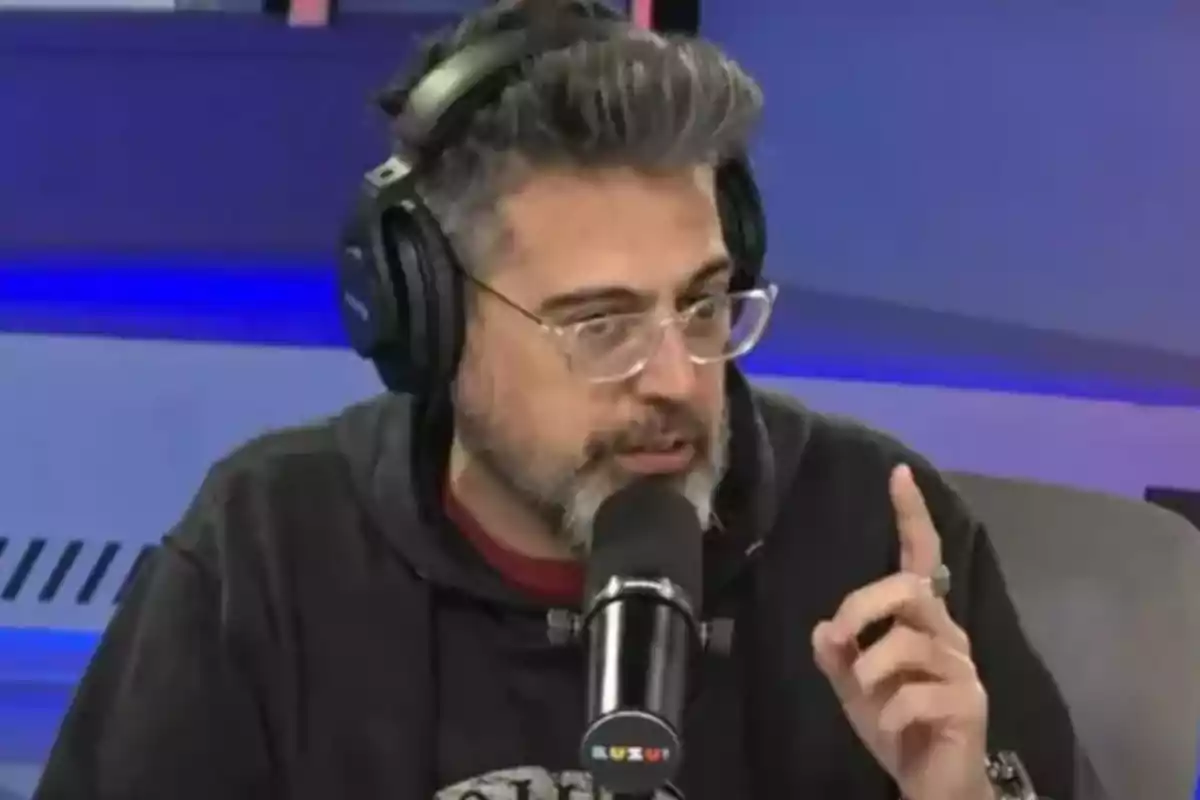 Hombre con gafas y audífonos hablando frente a un micrófono en un estudio de radio mientras levanta un dedo
