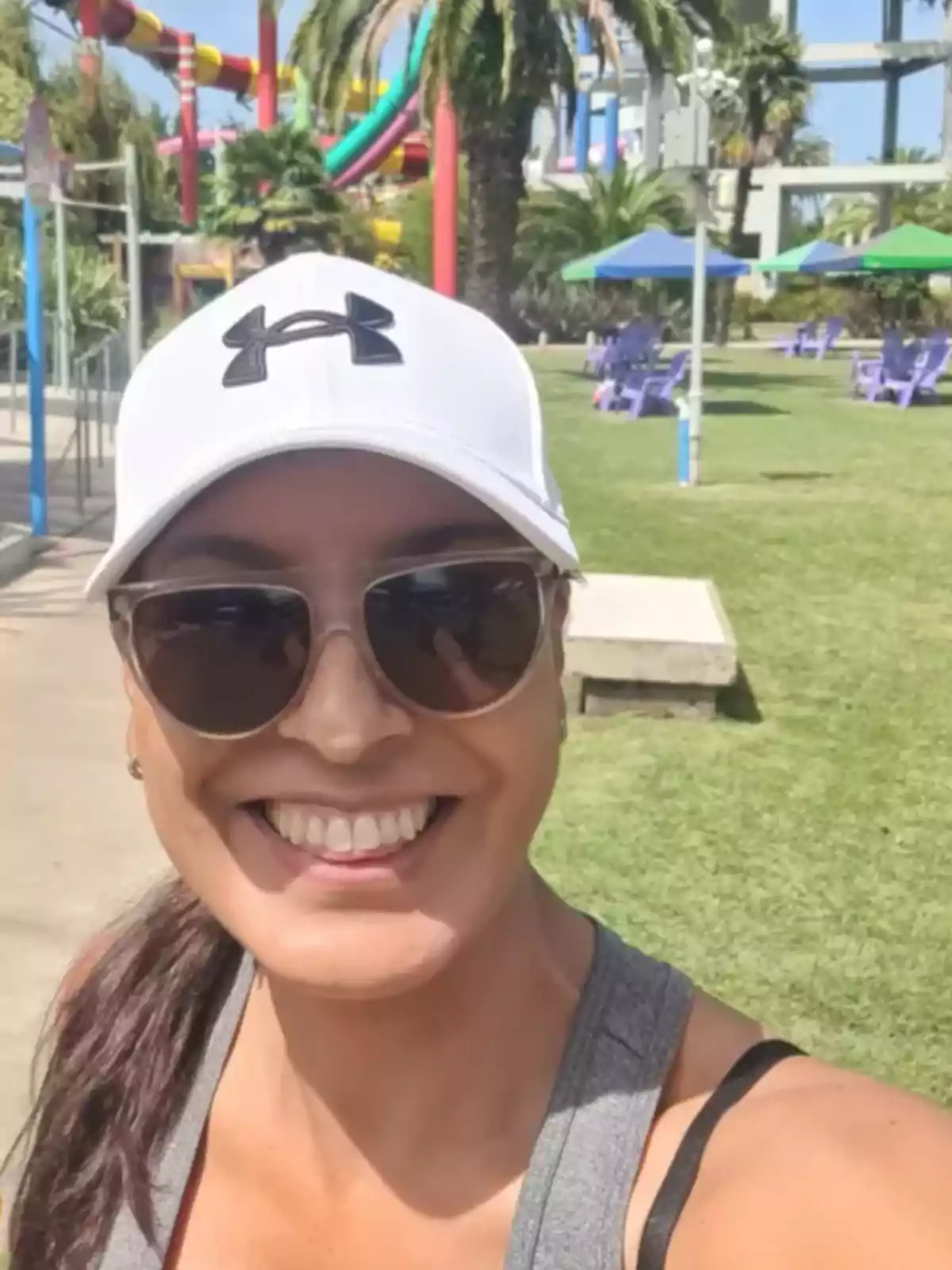 Smiling woman wearing a white cap and sunglasses taking a selfie at a water park with colorful slides and palm trees in the background Smiling woman wearing a white cap and sunglasses taking a selfie at a water park with colorful slides and palm trees in the background
