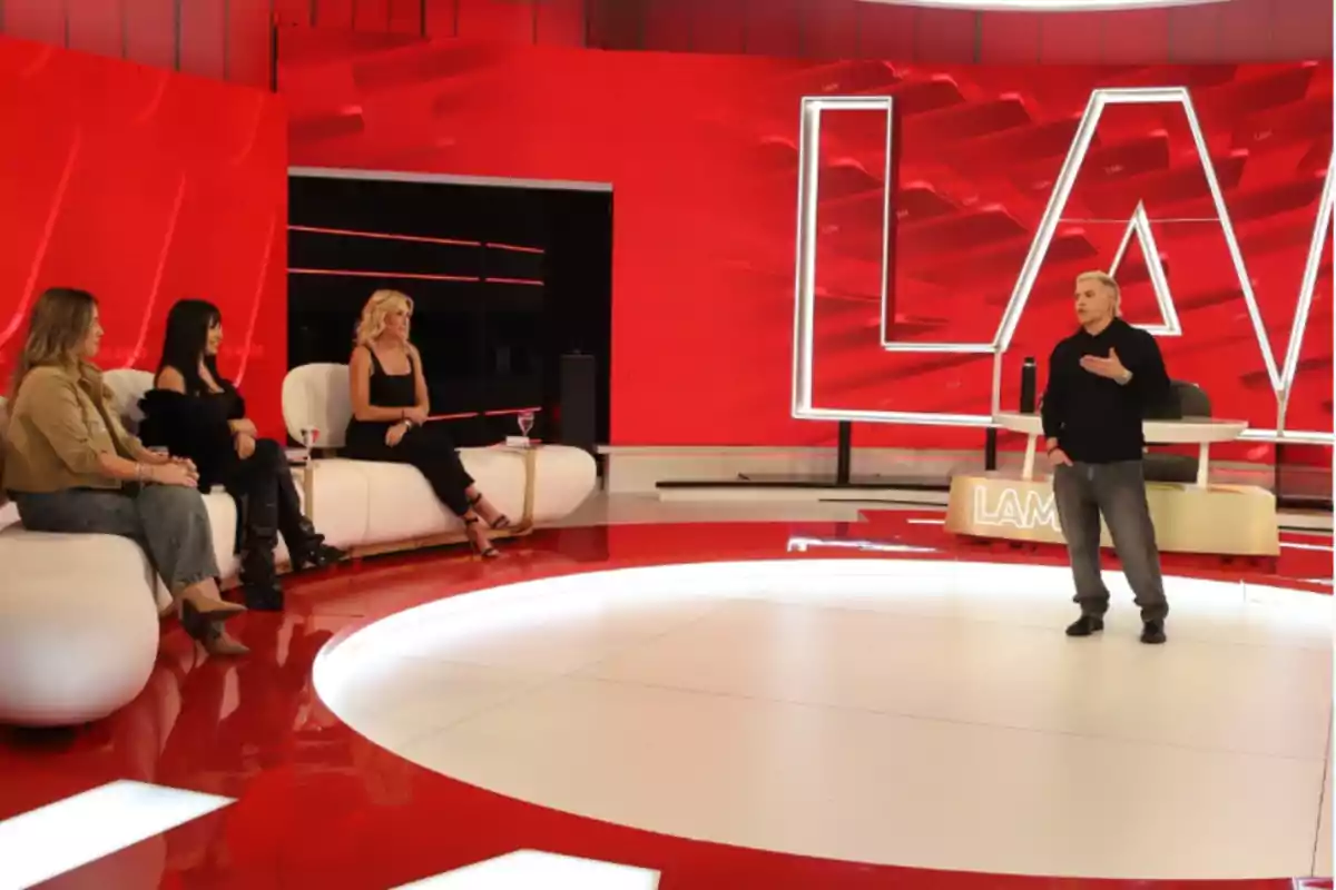A man standing speaks in front of three women seated on a modern television set with a red background and large illuminated letters. A man standing speaks in front of three women seated on a modern television set with a red background and large illuminated letters.