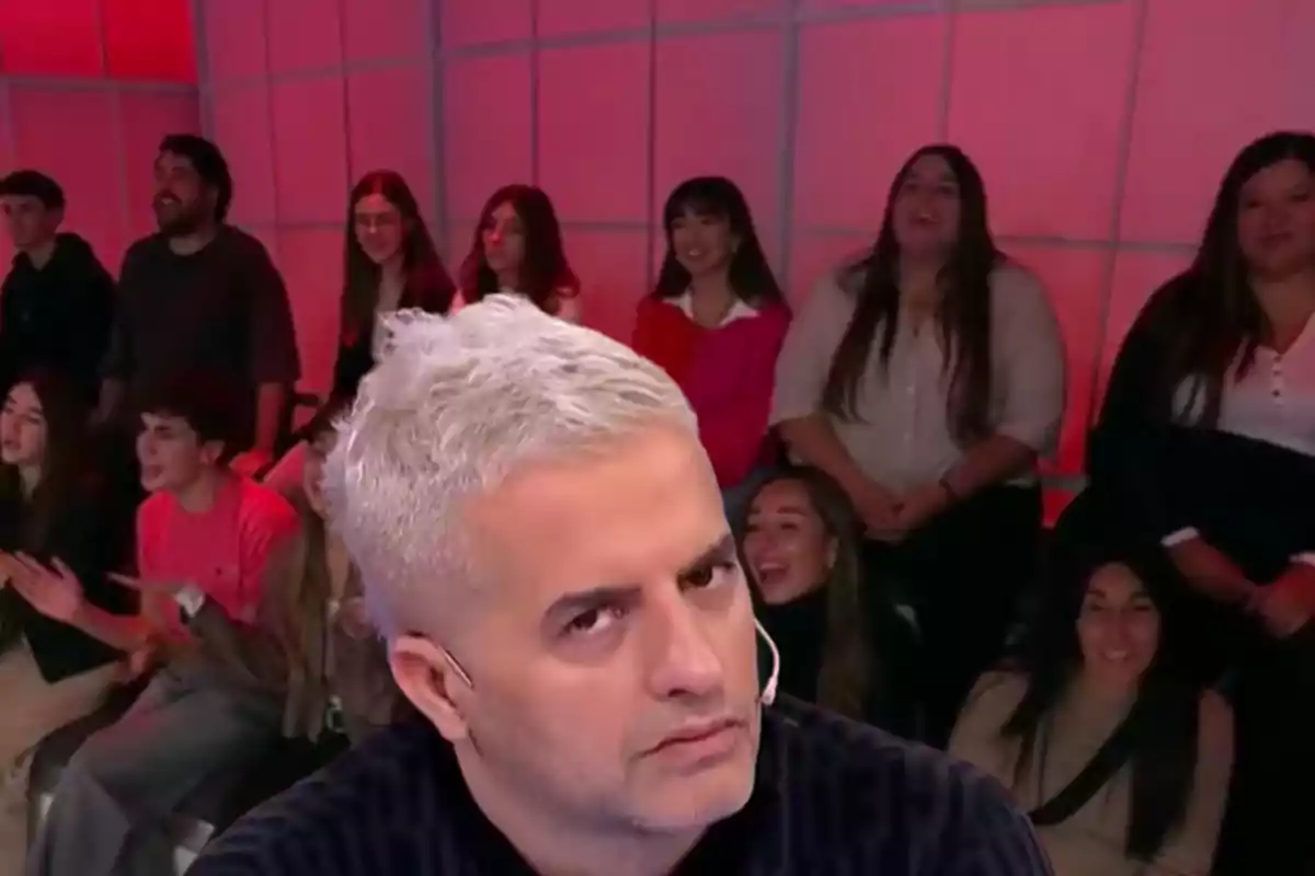 Gray-haired man with a serious expression in the foreground while several people smile and watch in the background on a set lit with red lights