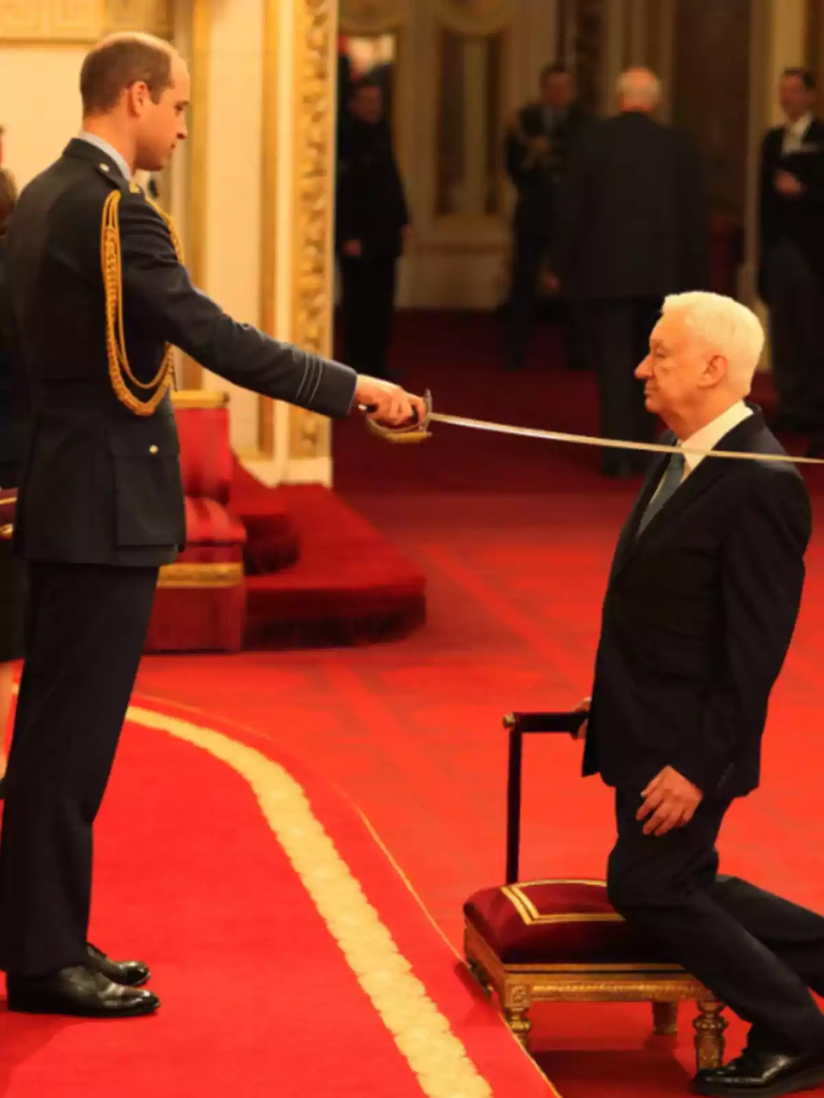 Un hombre de uniforme realiza una ceremonia de investidura con una espada a un hombre mayor arrodillado en un cojín en un salón elegante Un hombre de uniforme realiza una ceremonia de investidura con una espada a un hombre mayor arrodillado en un cojín en un salón elegante