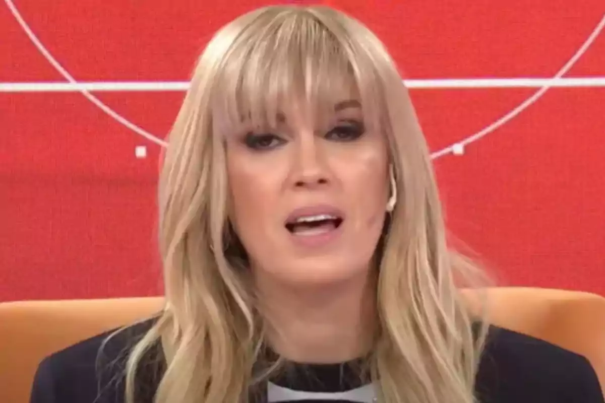 Mujer rubia con flequillo hablando en un set de televisión con fondo rojo