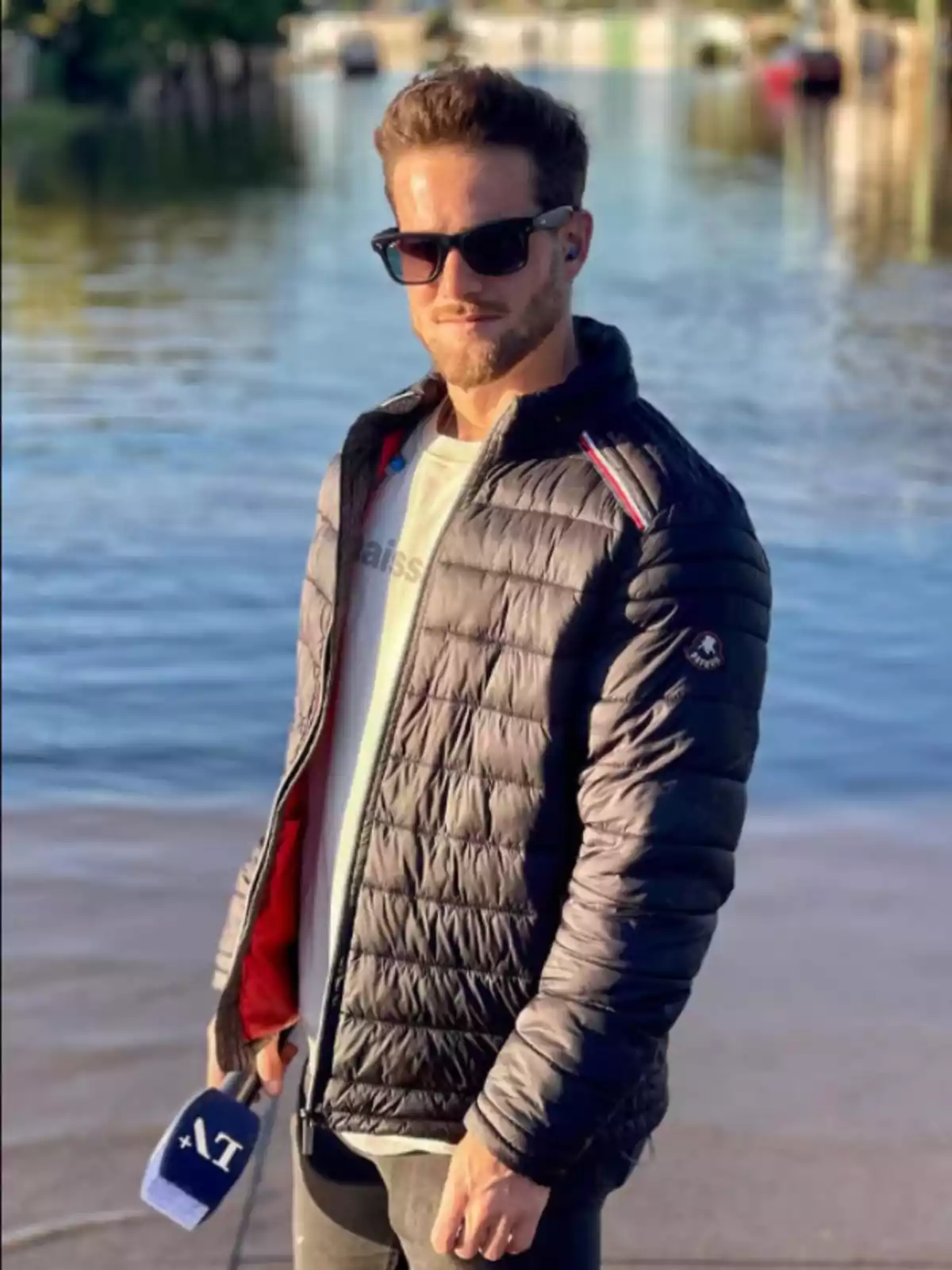 Man with sunglasses and a puffer jacket standing next to a body of water holding a blue microphone Man with sunglasses and a puffer jacket standing next to a body of water holding a blue microphone