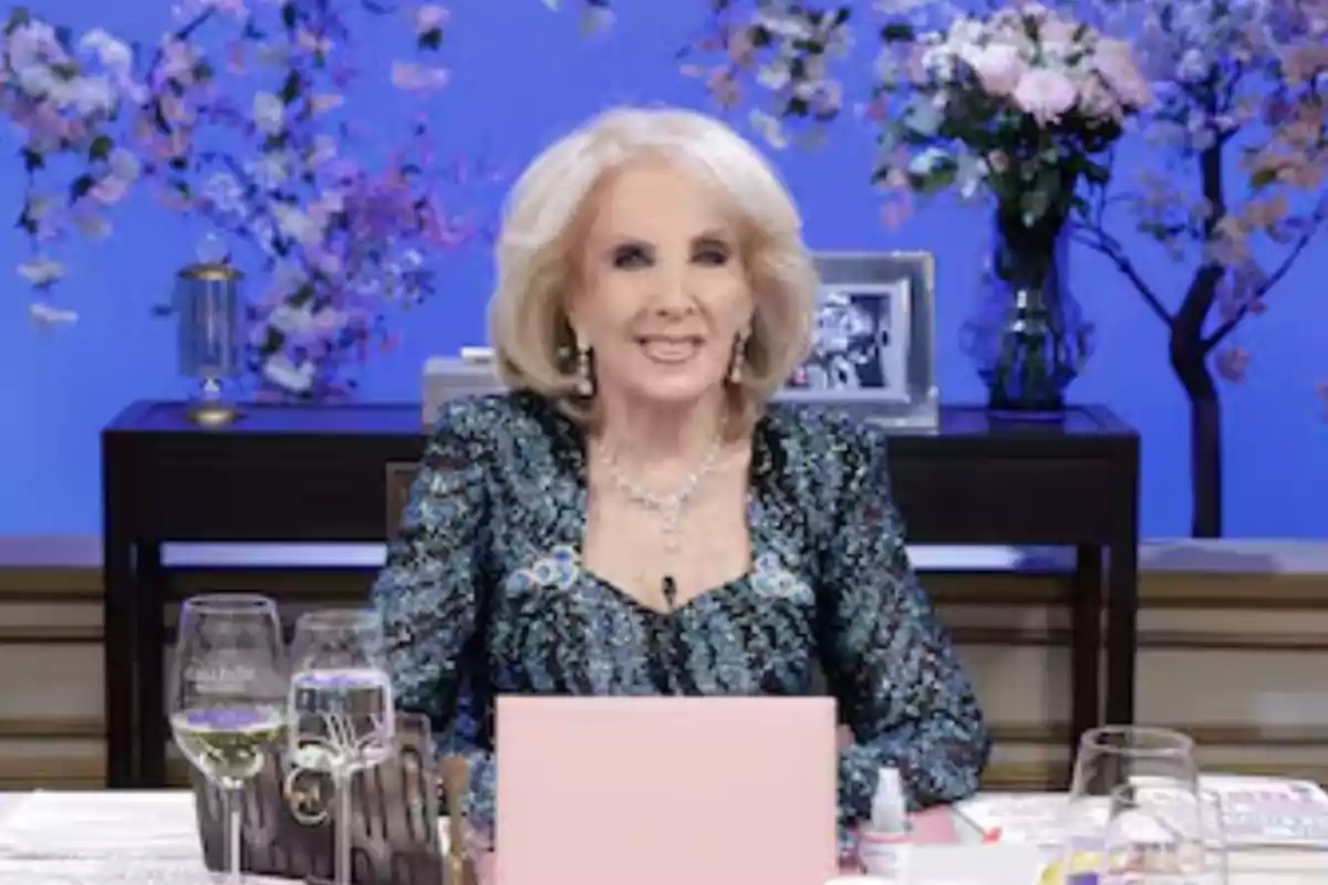 Older blonde woman sitting at an elegantly decorated table with glasses and flowers, blue background with floral arrangements and a black-and-white portrait behind her