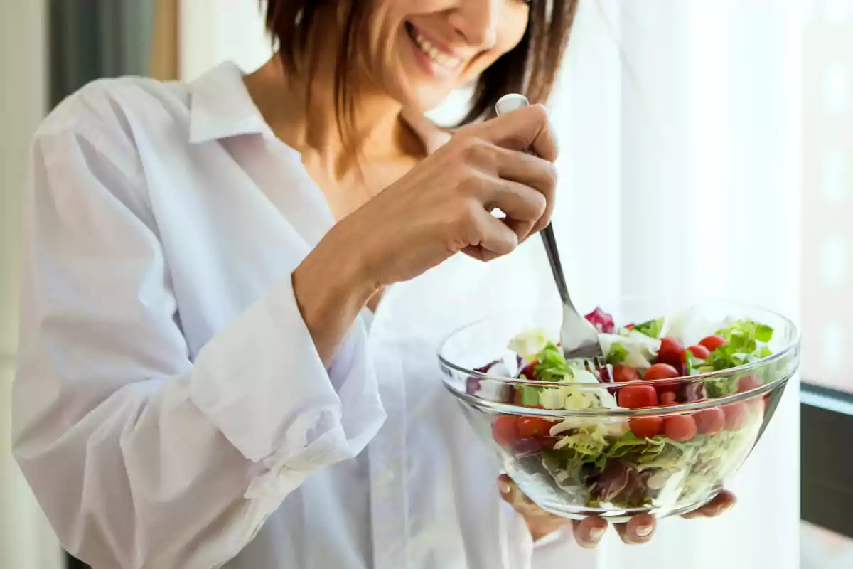 Mujer sonriente con camisa blanca sostiene un tazón de ensalada fresca con tomates y verduras mientras usa un tenedor Mujer sonriente con camisa blanca sostiene un tazón de ensalada fresca con tomates y verduras mientras usa un tenedor