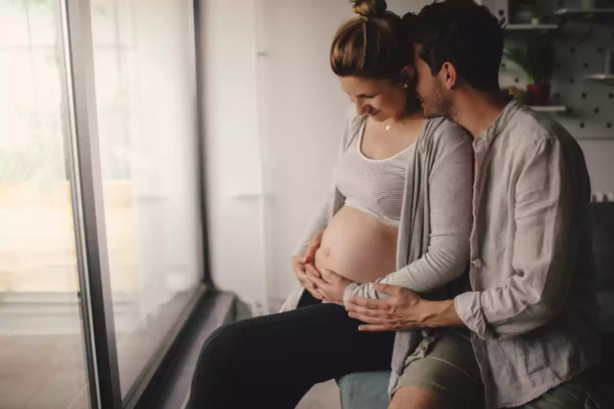 Pareja sentada junto a una ventana mientras el hombre abraza y acaricia la barriga de la mujer embarazada Pareja sentada junto a una ventana mientras el hombre abraza y acaricia la barriga de la mujer embarazada