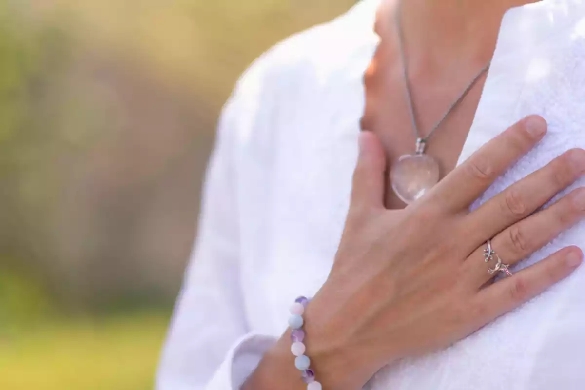 Persona con la mano sobre el pecho usando un collar de corazón y una pulsera de cuentas, vestida con ropa blanca Persona con la mano sobre el pecho usando un collar de corazón y una pulsera de cuentas, vestida con ropa blanca