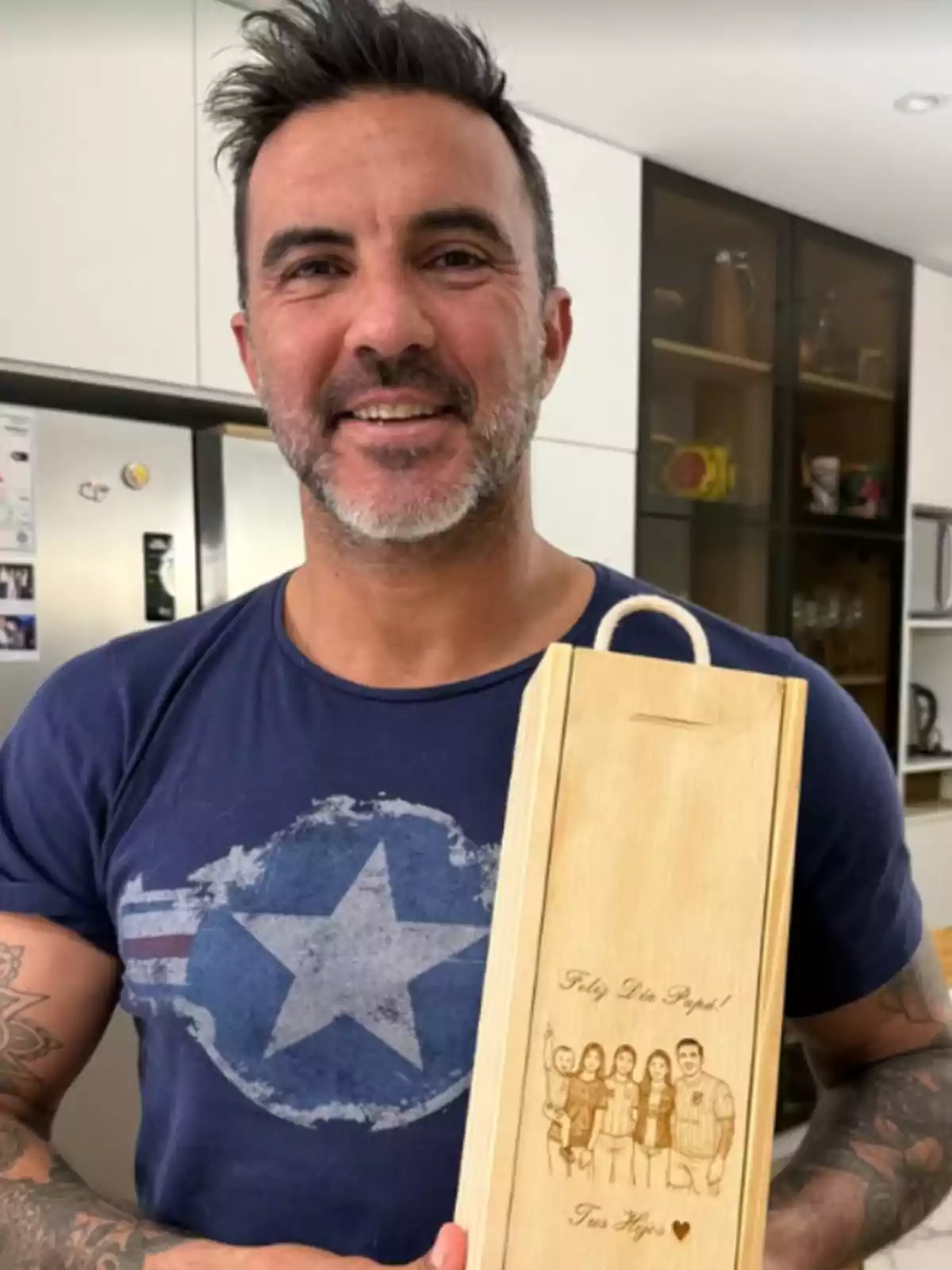 Hombre sonriente con barba y camiseta azul sostiene una caja de madera grabada con un dibujo familiar y un mensaje de felicitación en una cocina moderna. Hombre sonriente con barba y camiseta azul sostiene una caja de madera grabada con un dibujo familiar y un mensaje de felicitación en una cocina moderna.