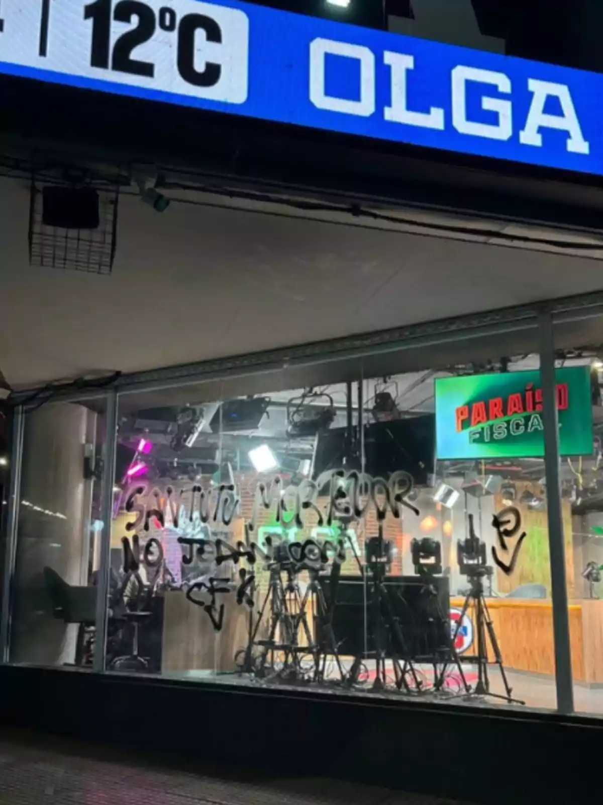 Estudio de radio con vidriera rayada con grafitis y luces encendidas en el interior, en la parte superior se ve un cartel digital que indica 12 grados y la palabra OLGA Estudio de radio con vidriera rayada con grafitis y luces encendidas en el interior, en la parte superior se ve un cartel digital que indica 12 grados y la palabra OLGA