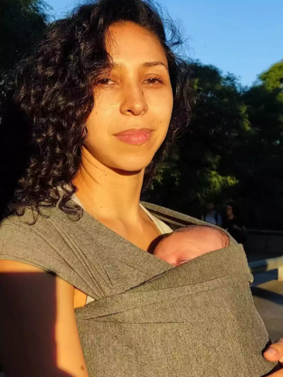 Curly-haired woman holds a baby in a gray fabric carrier while basking in the sunlight outdoors. Curly-haired woman holds a baby in a gray fabric carrier while basking in the sunlight outdoors.