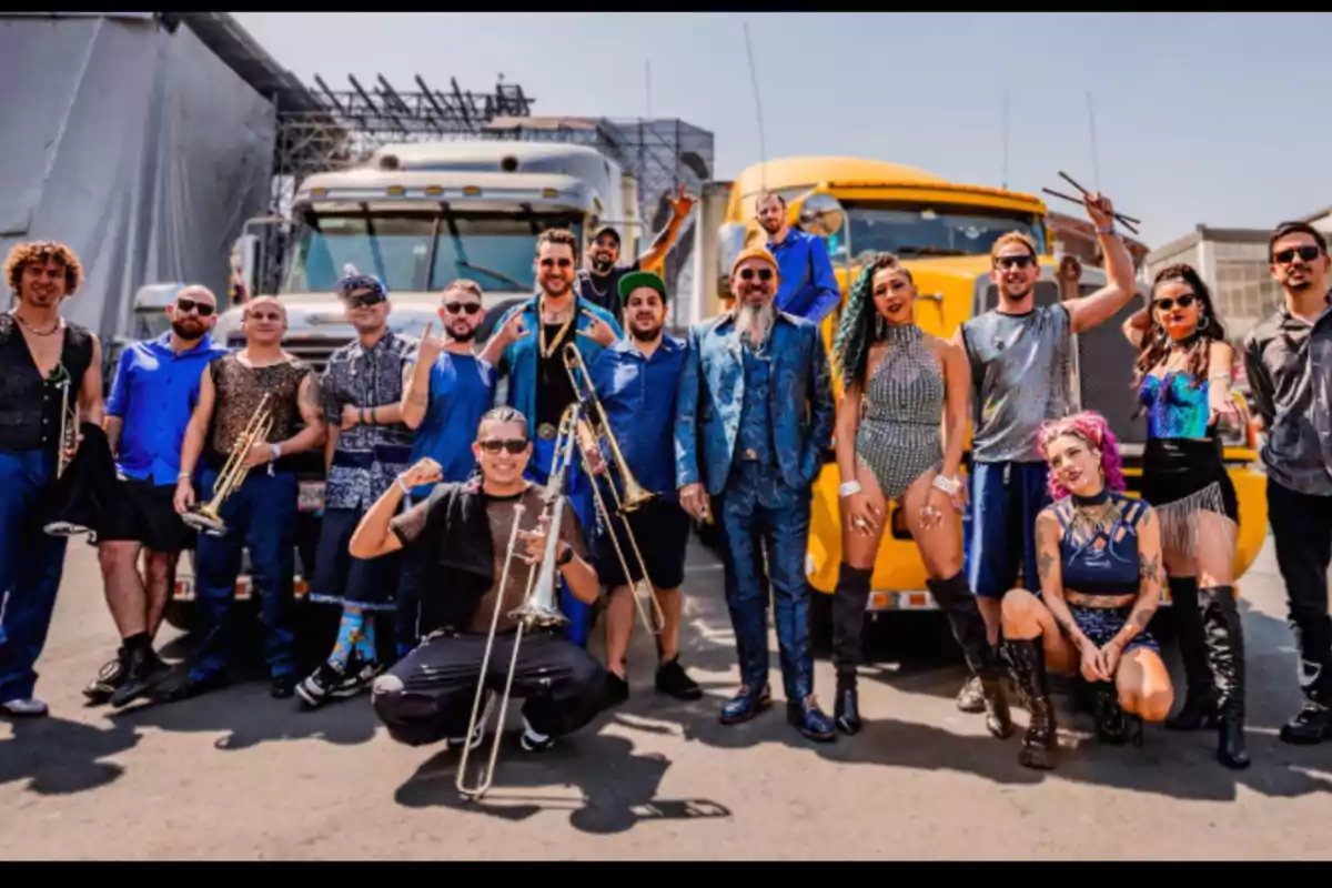 Group of musicians and dancers posing together outdoors in front of two large trucks, some holding brass instruments and all wearing striking, colorful costumes. Group of musicians and dancers posing together outdoors in front of two large trucks, some holding brass instruments and all wearing striking, colorful costumes.