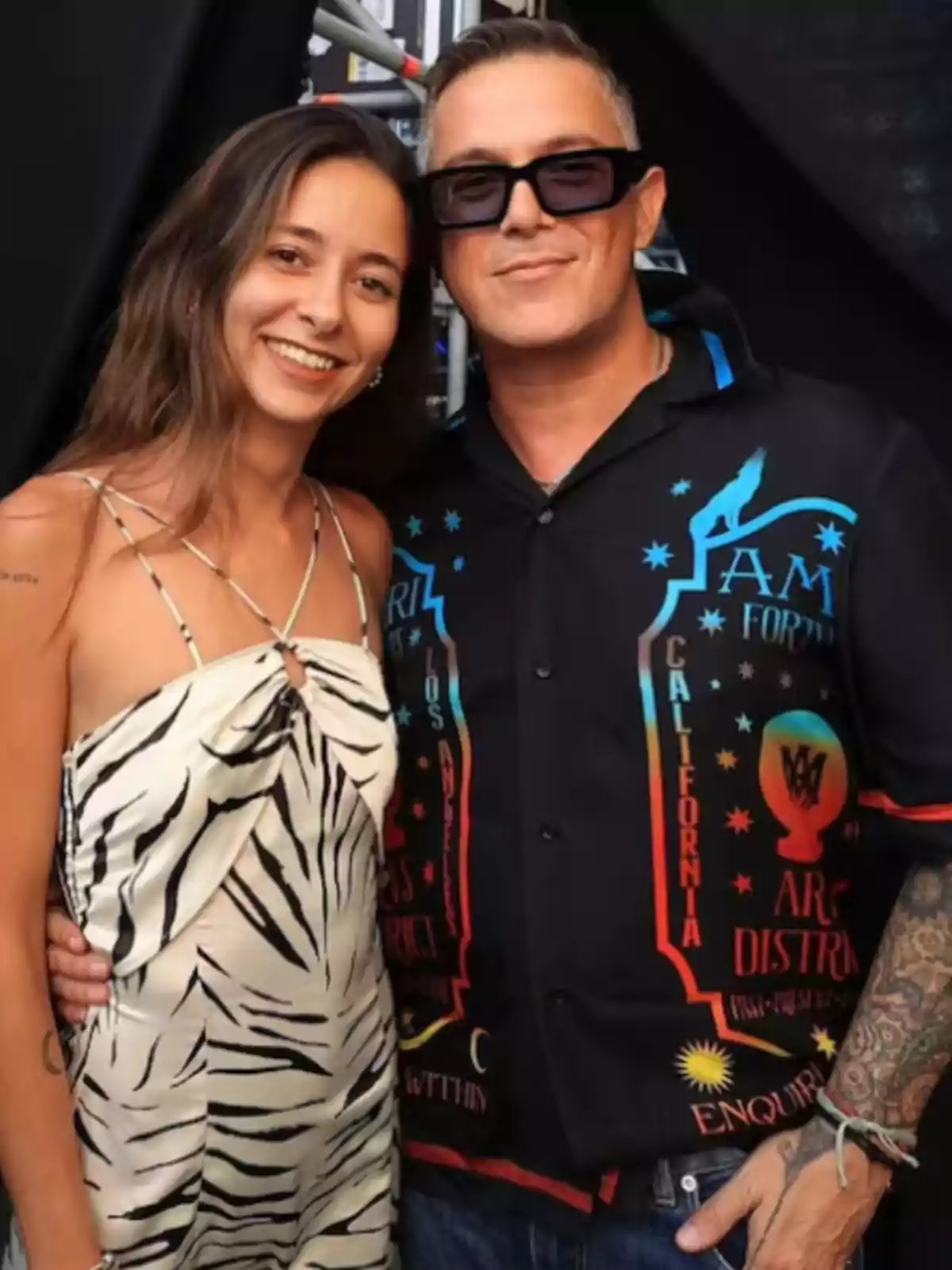 Una mujer con vestido de estampado animal y un hombre con camisa negra estampada y gafas oscuras posan juntos sonriendo frente a la cámara. Una mujer con vestido de estampado animal y un hombre con camisa negra estampada y gafas oscuras posan juntos sonriendo frente a la cámara.