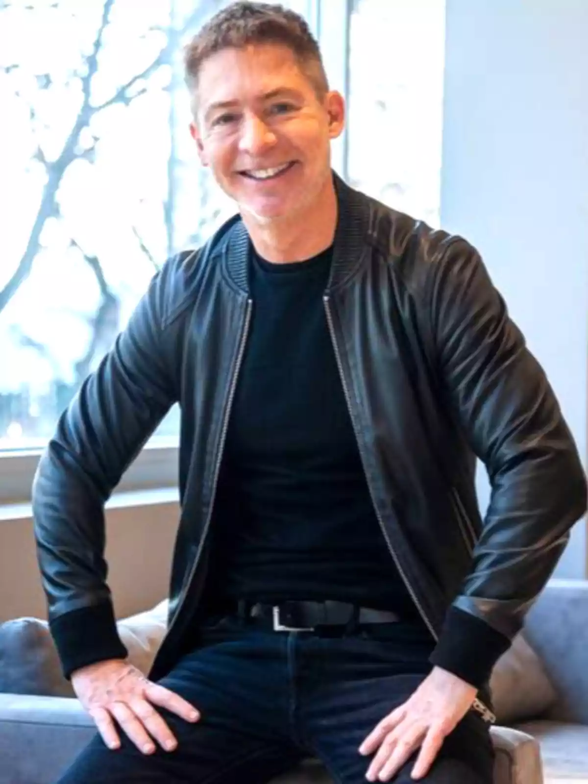Hombre sonriente con chaqueta de cuero negra y camiseta oscura sentado en un sillón junto a una ventana Hombre sonriente con chaqueta de cuero negra y camiseta oscura sentado en un sillón junto a una ventana