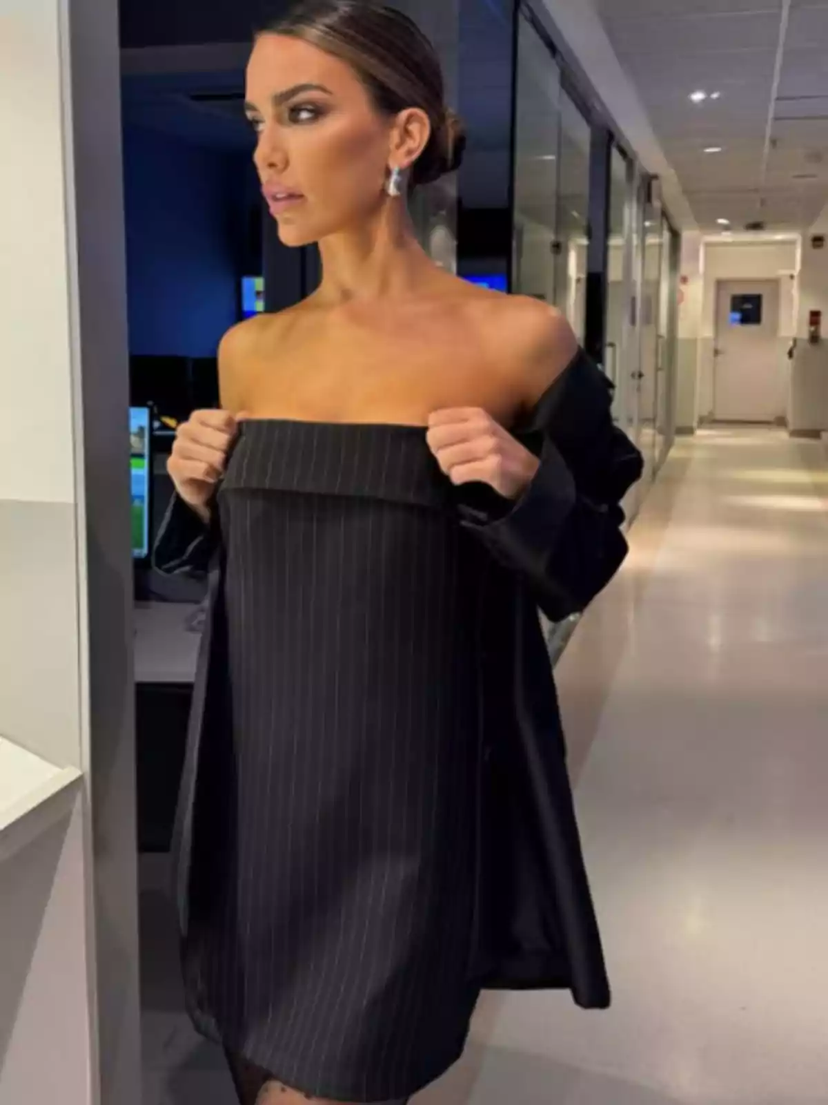 Mujer con vestido negro de rayas y saco a juego, de pie en un pasillo iluminado de oficina, mirando hacia un lado Mujer con vestido negro de rayas y saco a juego, de pie en un pasillo iluminado de oficina, mirando hacia un lado