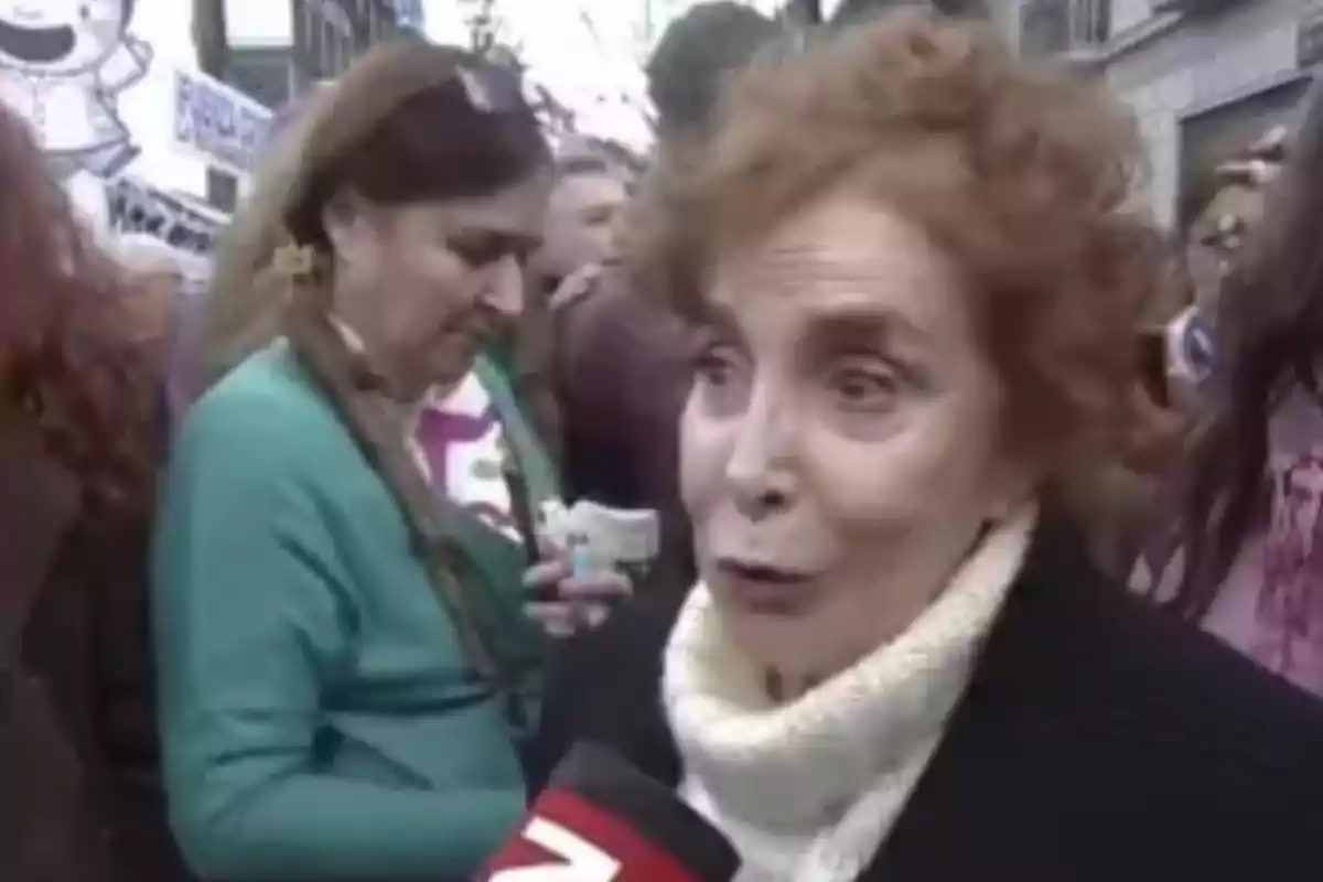 An older woman with curly hair and a white scarf is being interviewed on the street during a protest, while other people can be seen in the background. An older woman with curly hair and a white scarf is being interviewed on the street during a protest, while other people can be seen in the background.