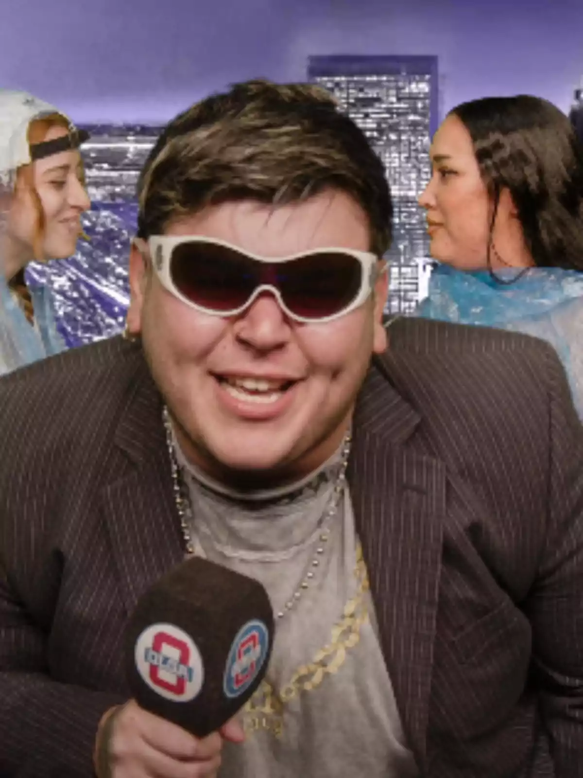 A man wearing sunglasses and holding a microphone smiles at the camera while two women talk in the background with a blurred urban landscape. A man wearing sunglasses and holding a microphone smiles at the camera while two women talk in the background with a blurred urban landscape.