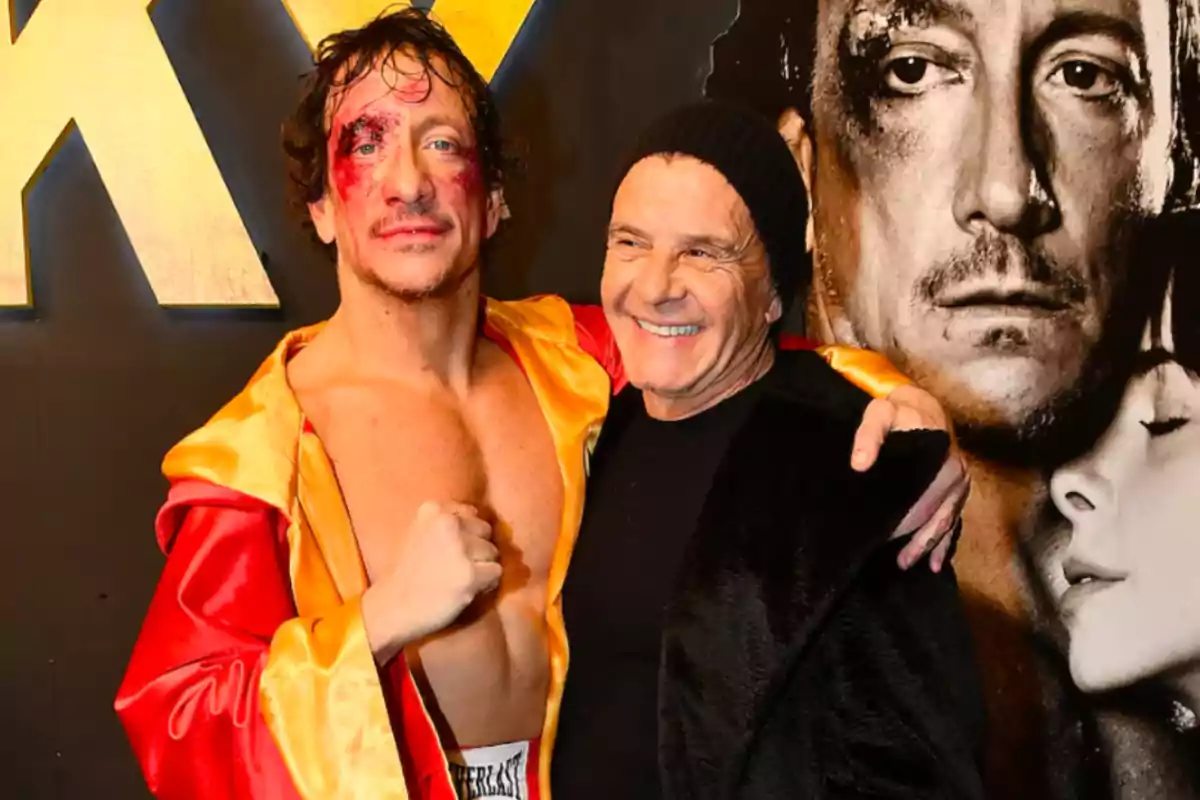 Two men pose together in front of a sign; one of them is wearing a boxing robe and has fake wounds on his face, while the other smiles and hugs him. Two men pose together in front of a sign; one of them is wearing a boxing robe and has fake wounds on his face, while the other smiles and hugs him.