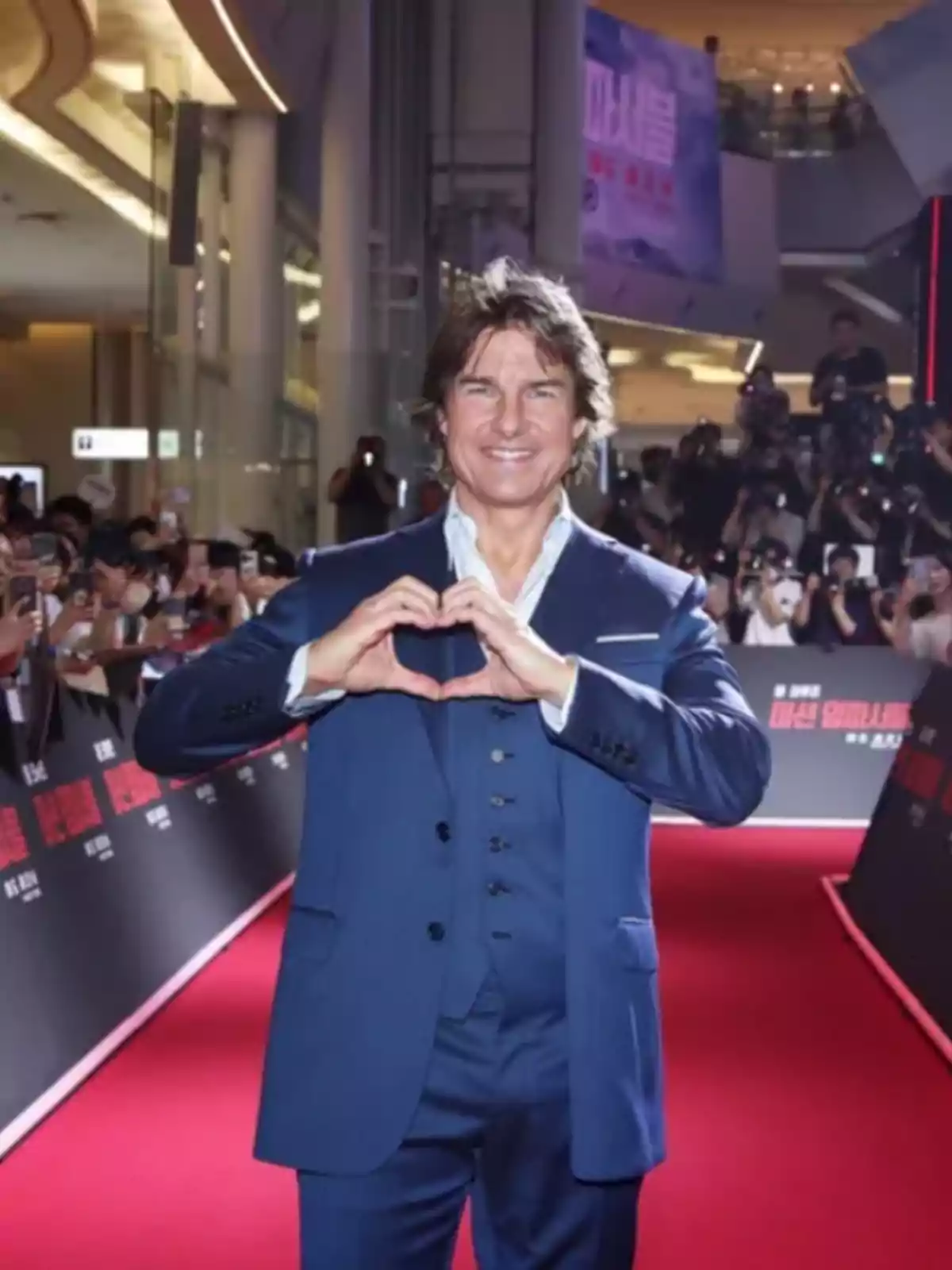 Hombre con traje azul sonríe y hace un gesto de corazón con las manos en una alfombra roja rodeado de fotógrafos y público. Hombre con traje azul sonríe y hace un gesto de corazón con las manos en una alfombra roja rodeado de fotógrafos y público.